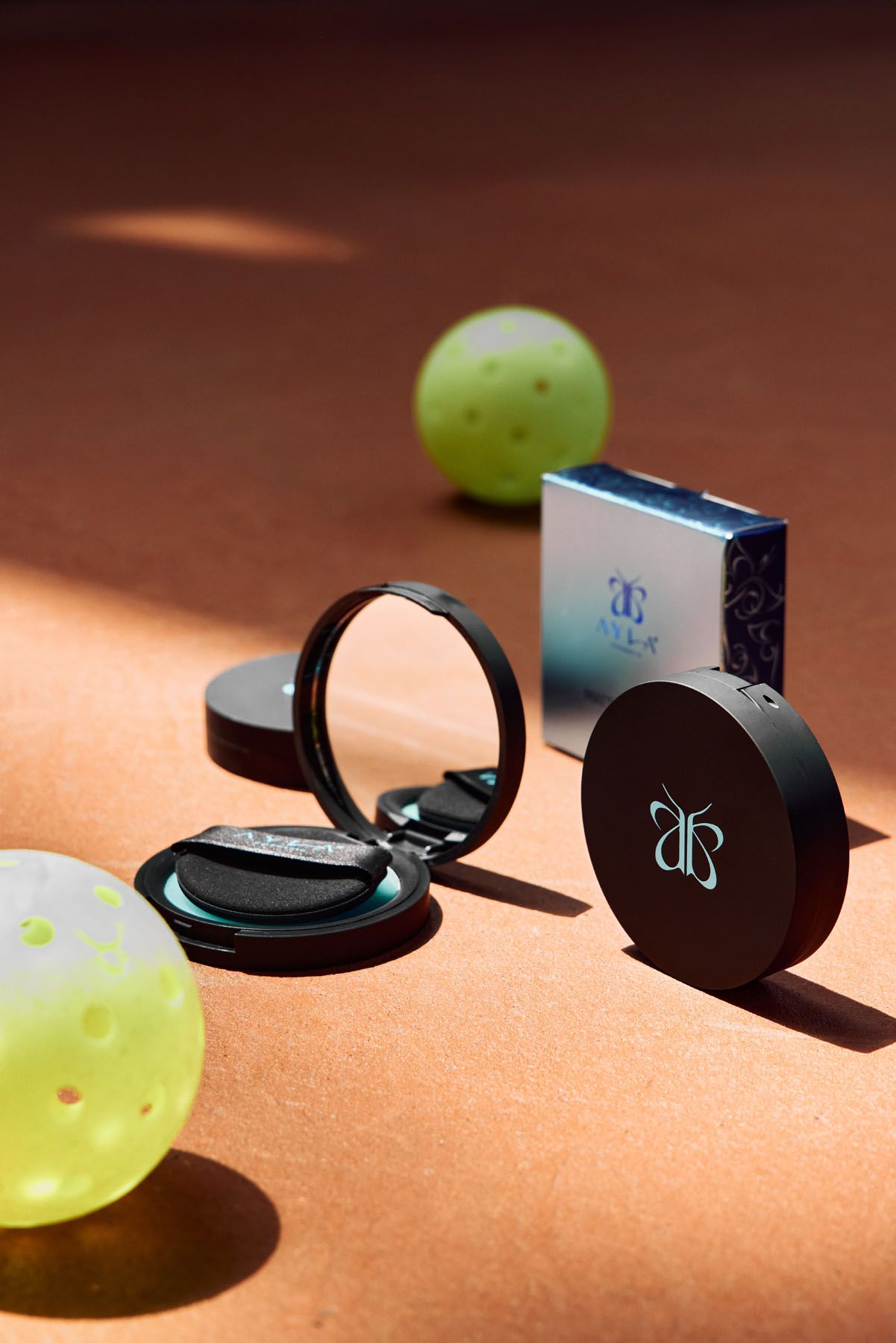 Anastasia Beverly Hills compact powder with mirror open, surrounded by yellow pickleball balls on a clay court surface.