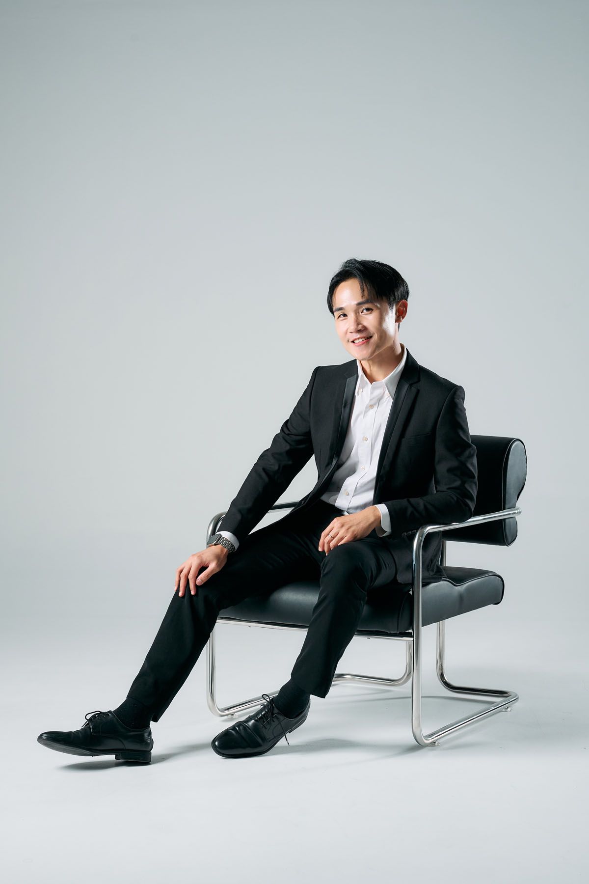 Smiling young Asian man in black suit sitting relaxed on leather chair against light gray background.