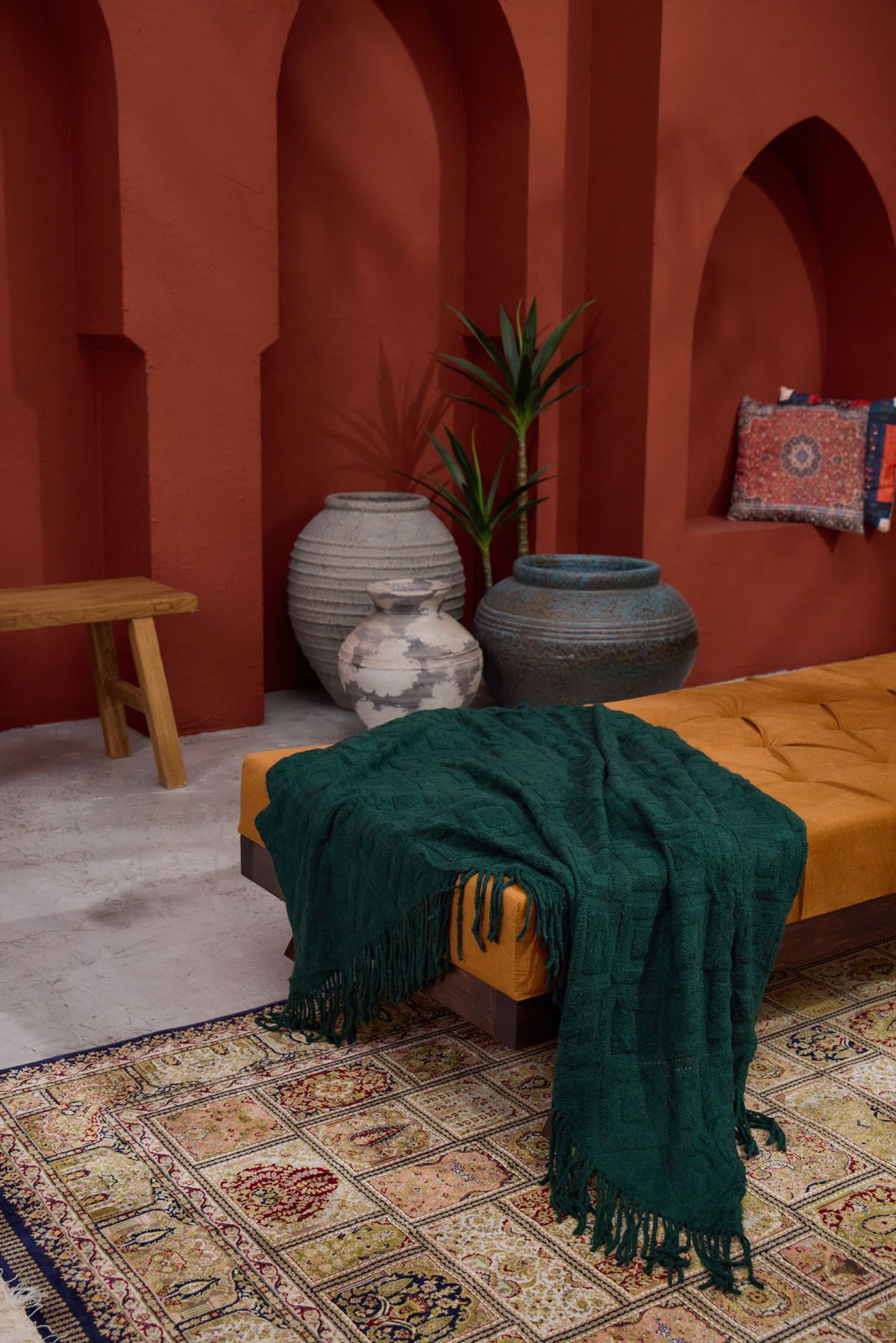 Emerald green fringed throw draped over mustard daybed in Moroccan-inspired terracotta room with ceramic vases.