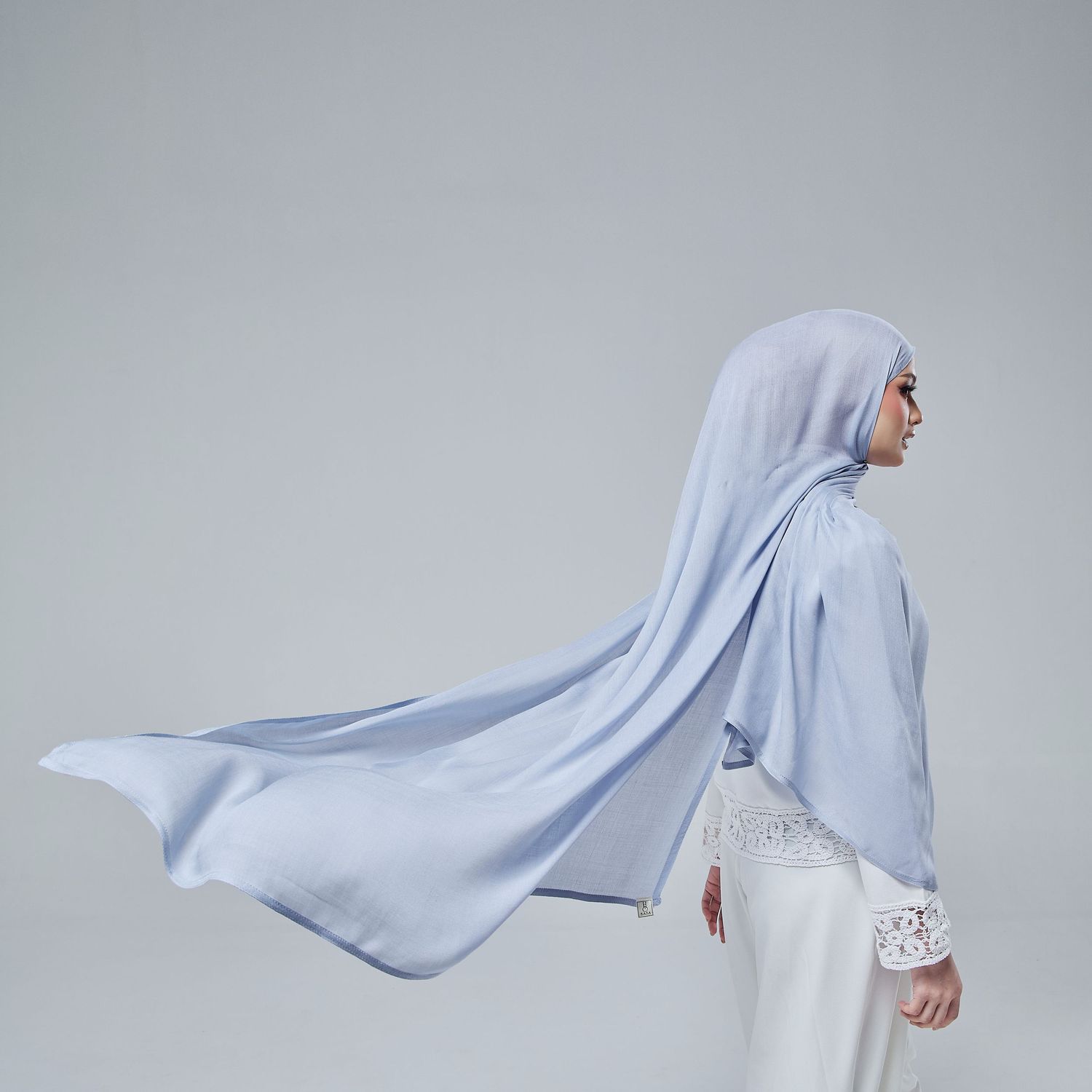 Woman wearing a flowing light blue hijab and white lace dress, posed elegantly against a neutral background.