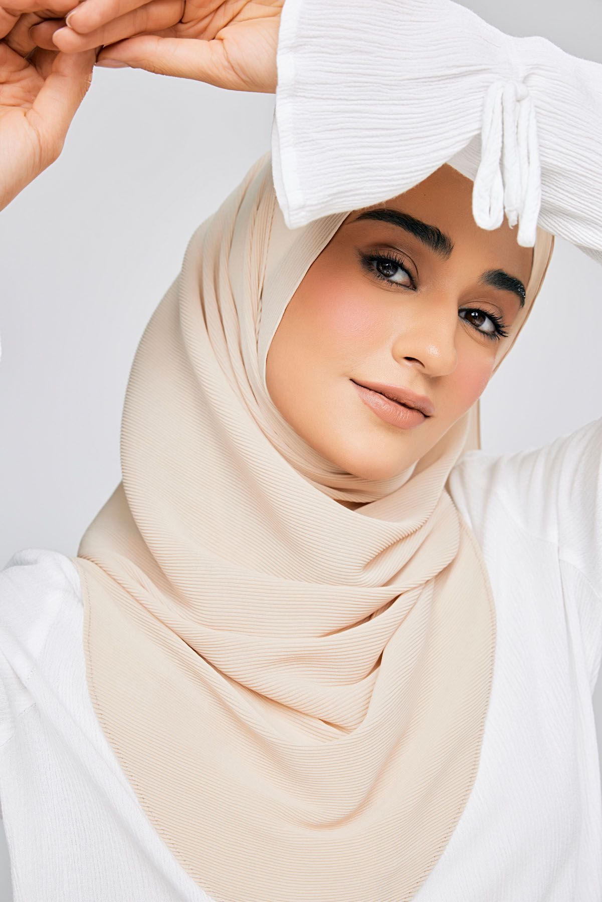 Woman wearing a beige hijab scarf and white top poses gracefully against a light gray background.