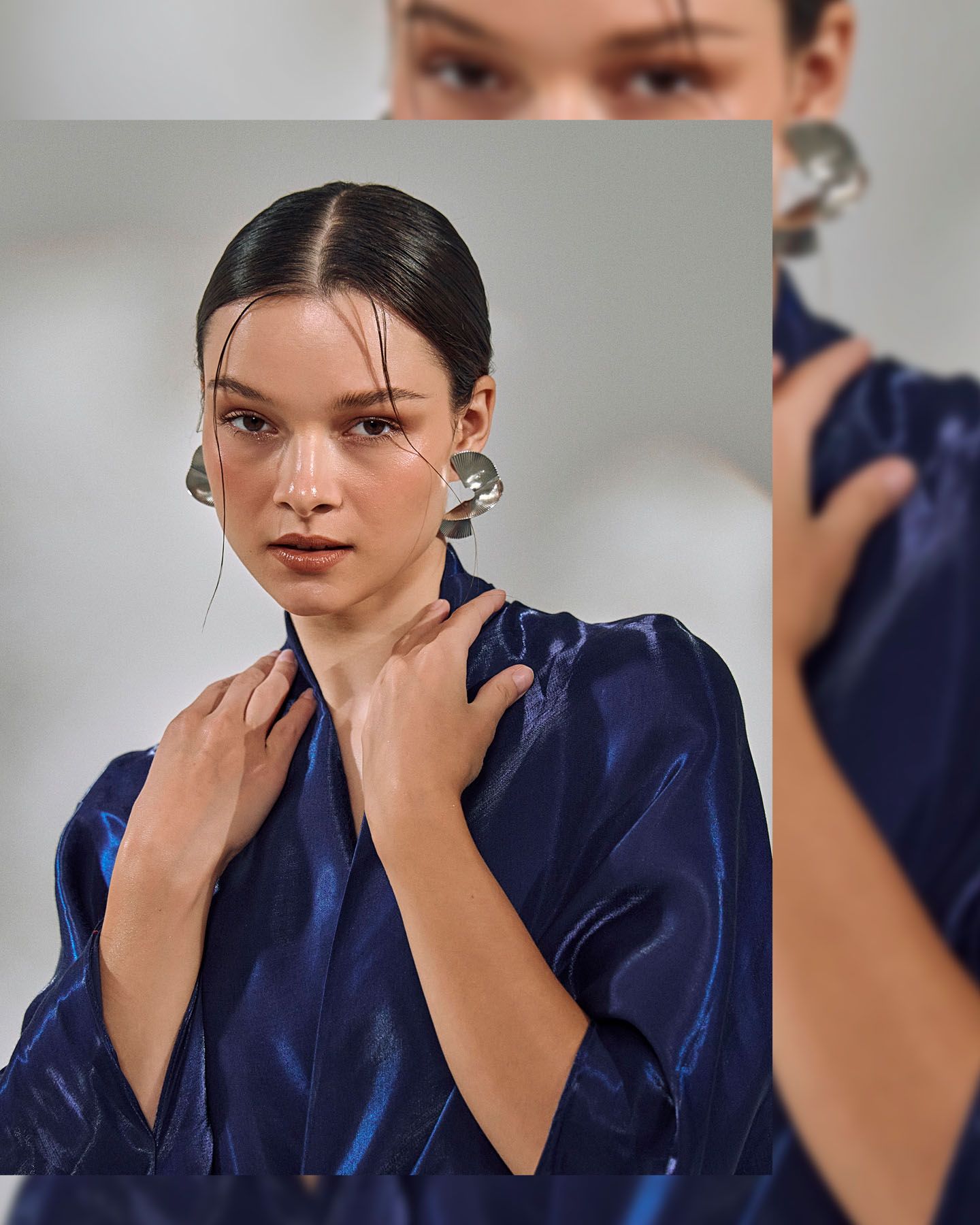 Model in blue satin robe with large silver earrings poses near mirror reflection on light background.