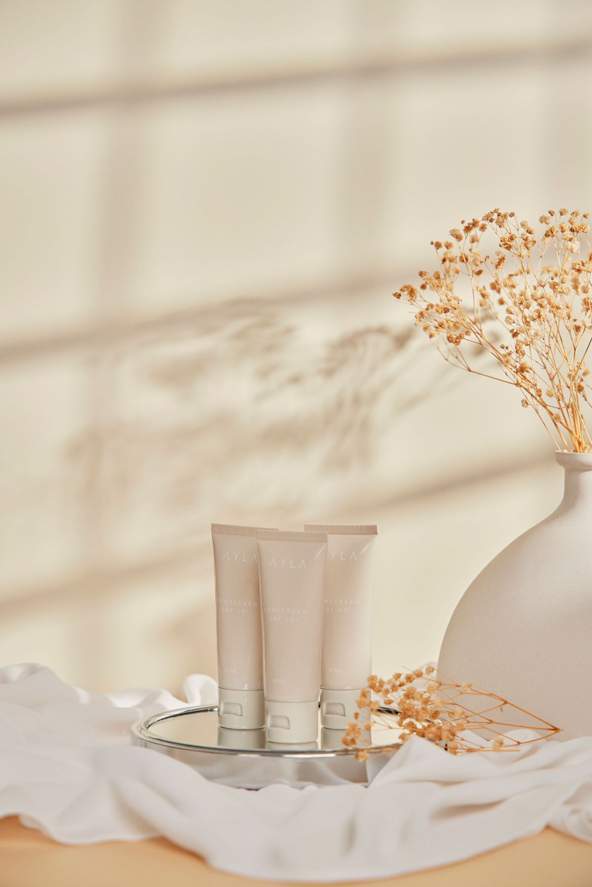 Three beige skincare cream tubes displayed on mirror tray with dried flowers and white fabric backdrop.