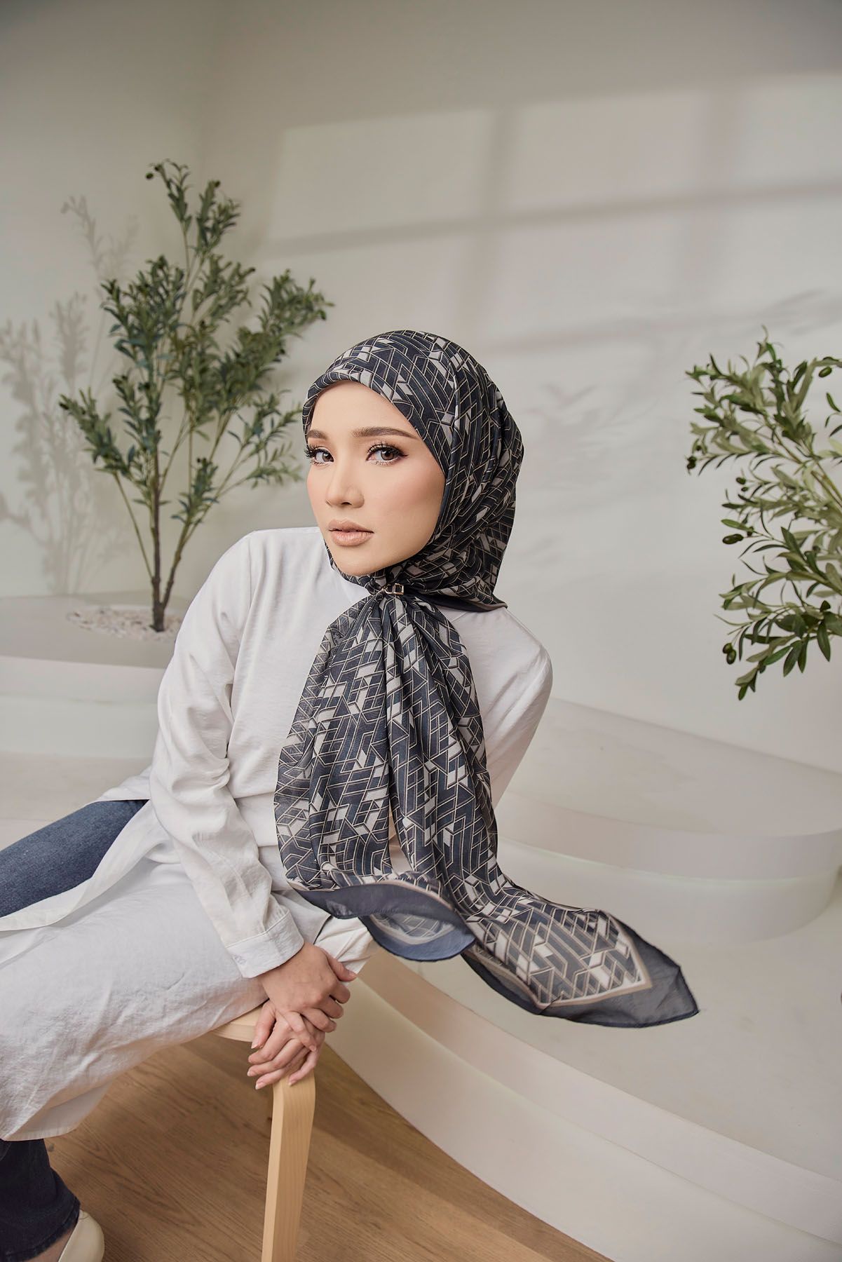 Woman in a dark patterned hijab and white outfit sitting on a bench near green plants.