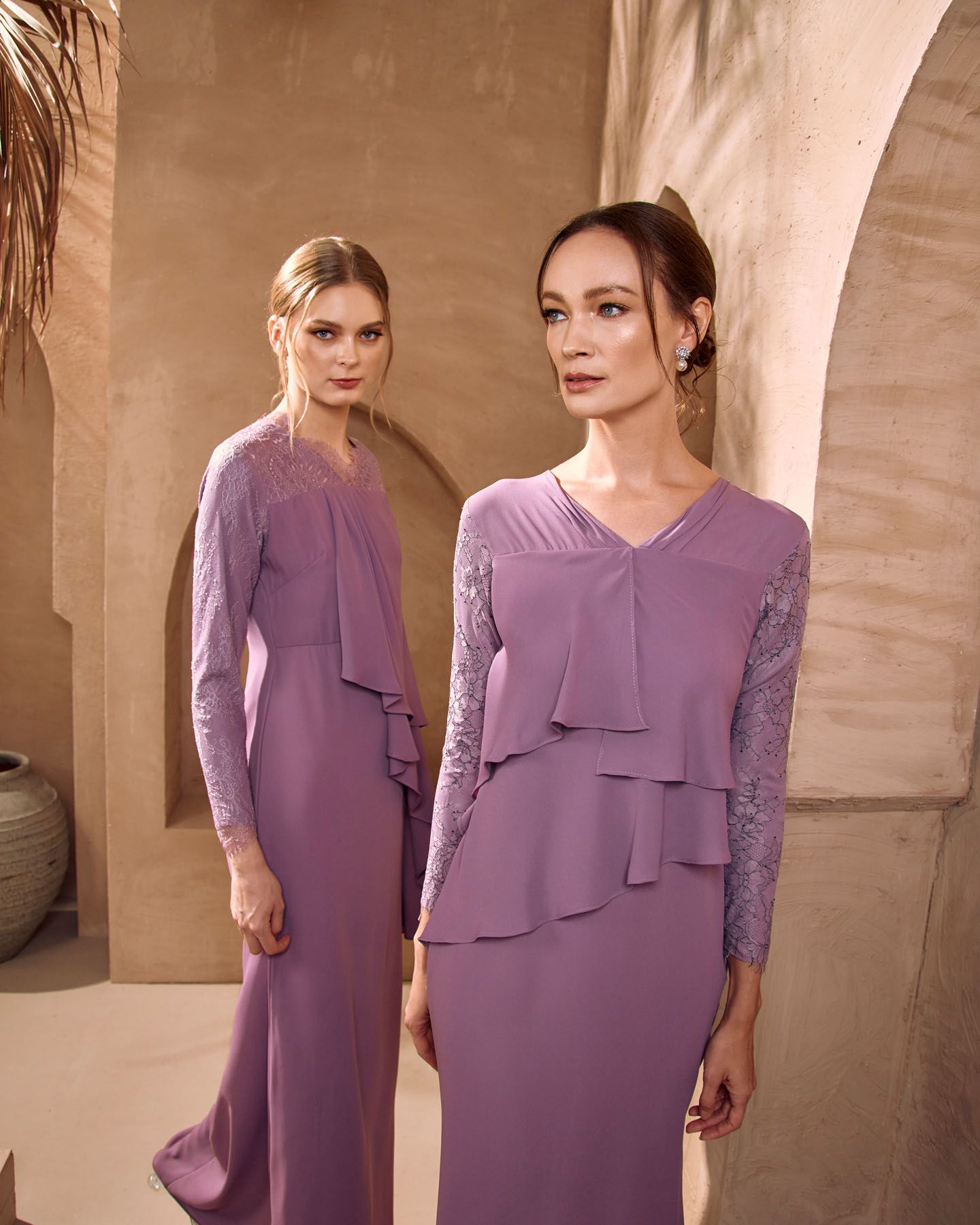 Two women modeling matching lavender purple modest dresses with lace sleeves in sandy archway.