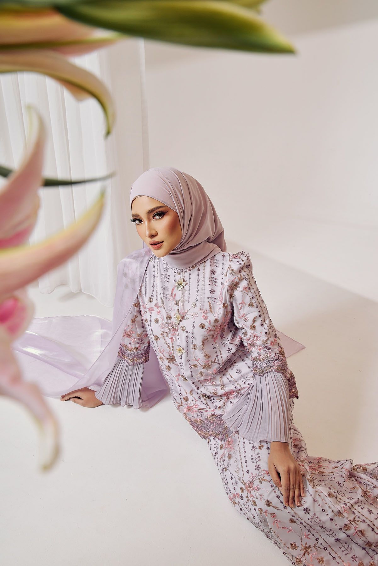 Muslim woman in pink floral modest dress and mauve hijab posing elegantly in a bright white studio setting.