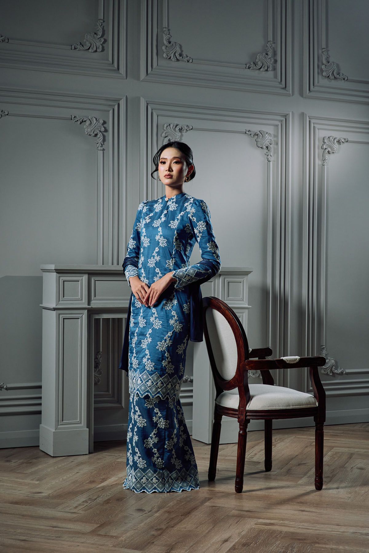 Woman in elegant blue lace kebaya gown stands beside a vintage chair in a sophisticated white-paneled room.