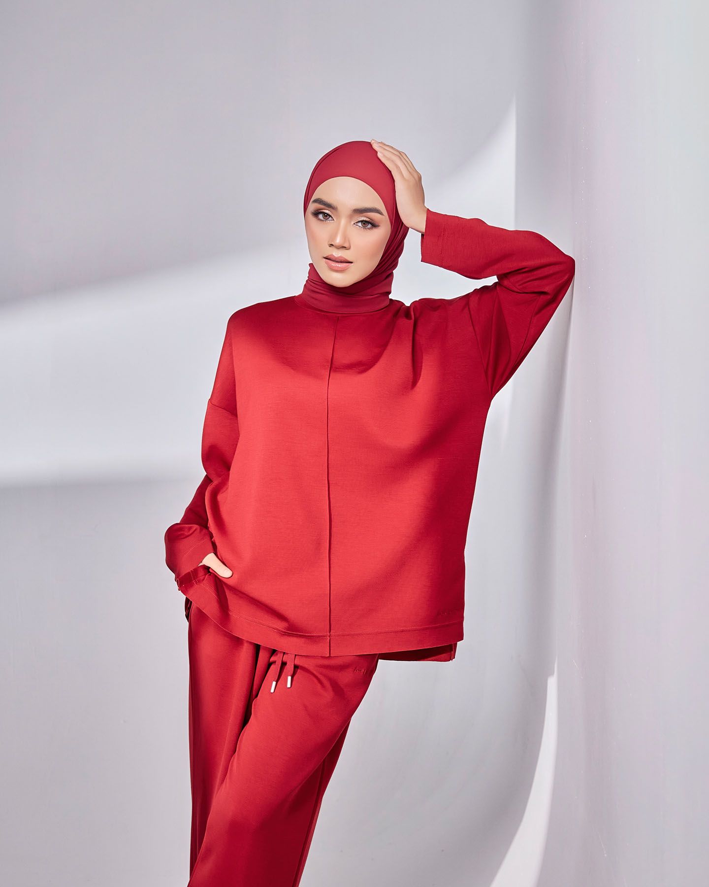 A woman in a matching red hijab and tracksuit set poses confidently against a white studio wall.