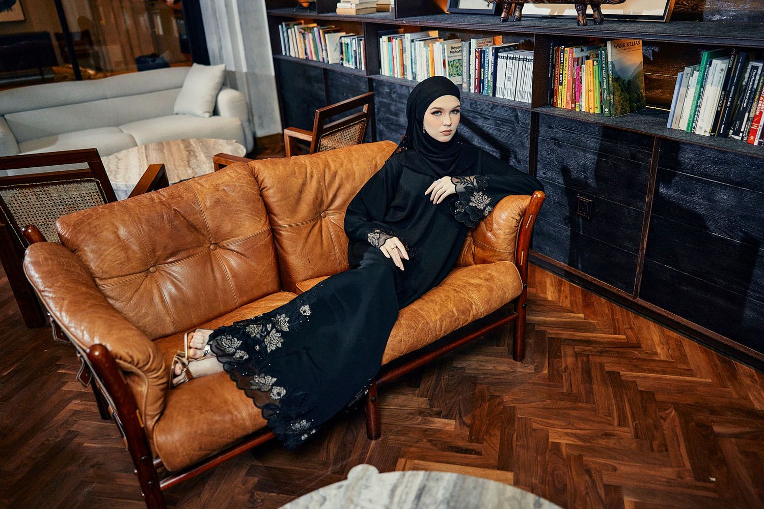 Muslim woman in black floral embroidered abaya and hijab reclining on brown leather sofa indoors.
