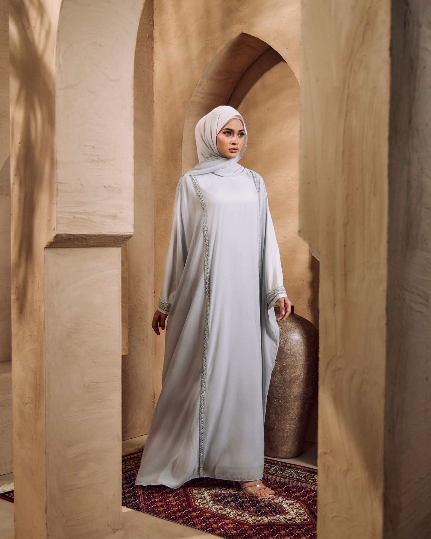 Woman in light grey abaya and matching hijab standing in arched doorway with terracotta walls.