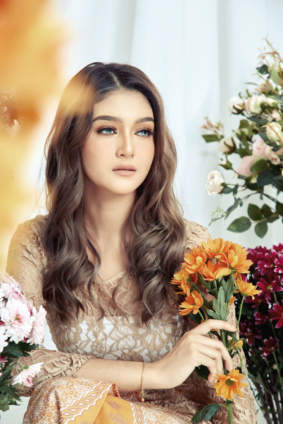 A beautiful young woman with long wavy hair holds orange flowers in an elegant lace dress.