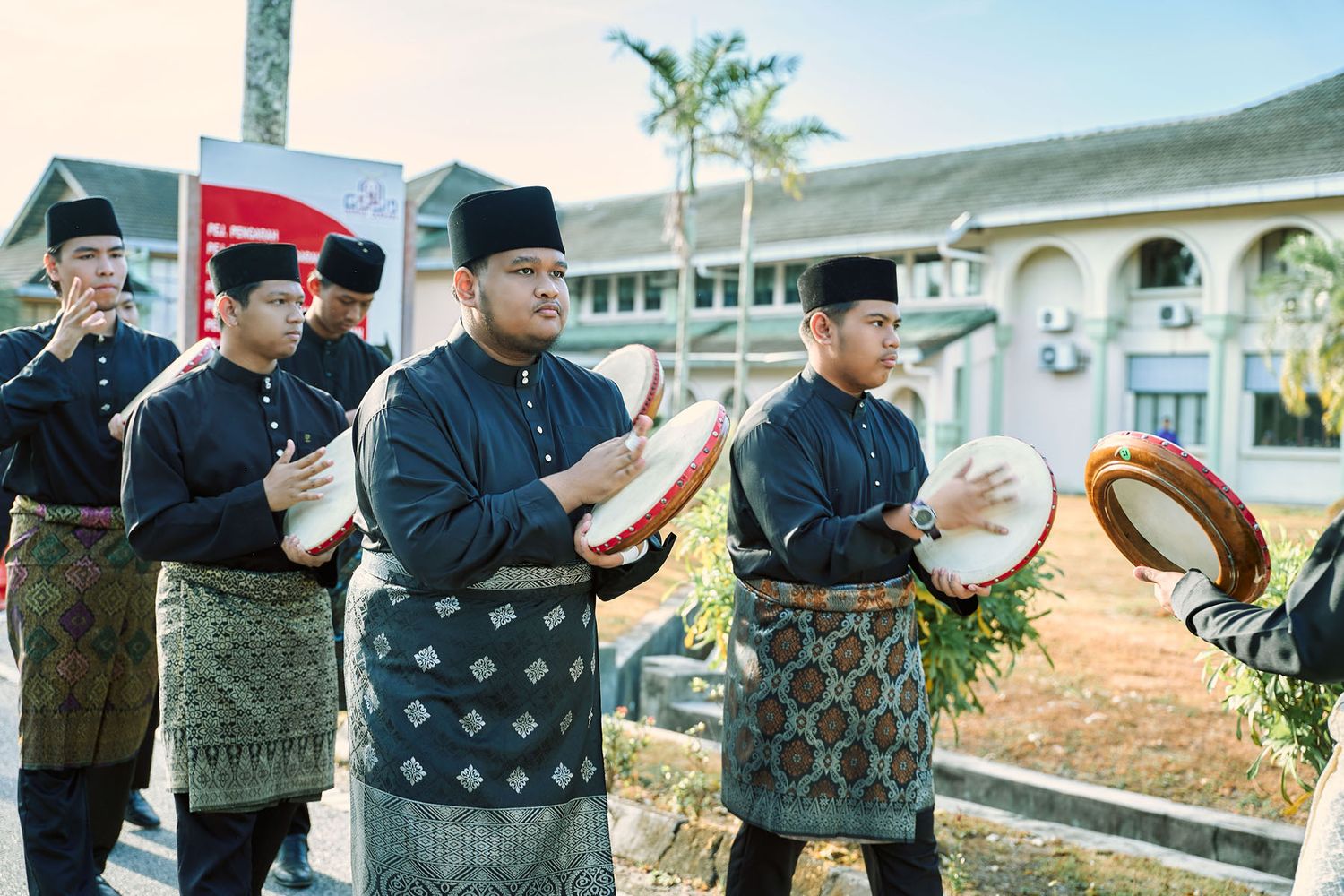Malaysian men in traditional black Baju Melayu playing kompang drums at an outdoor cultural ceremony.