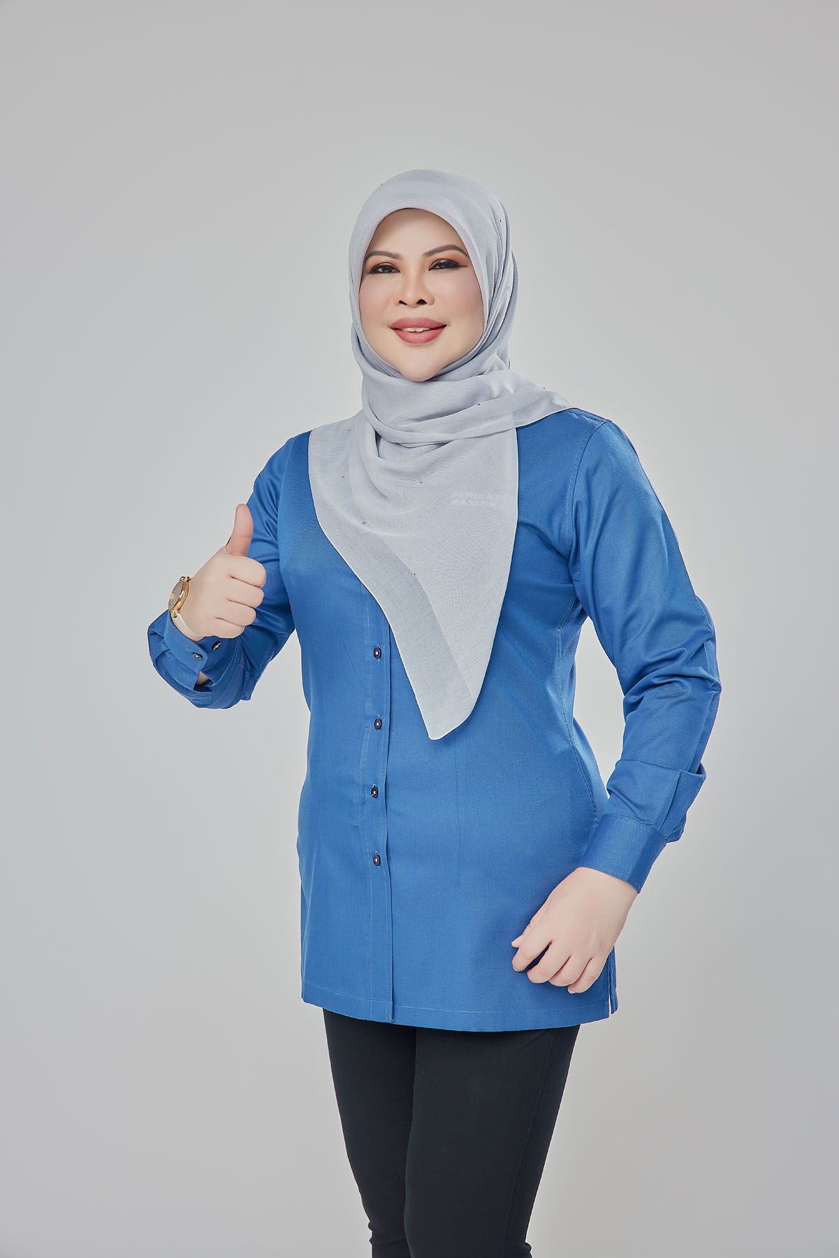 Muslim woman in blue button-up tunic and grey hijab giving thumbs up on white background.