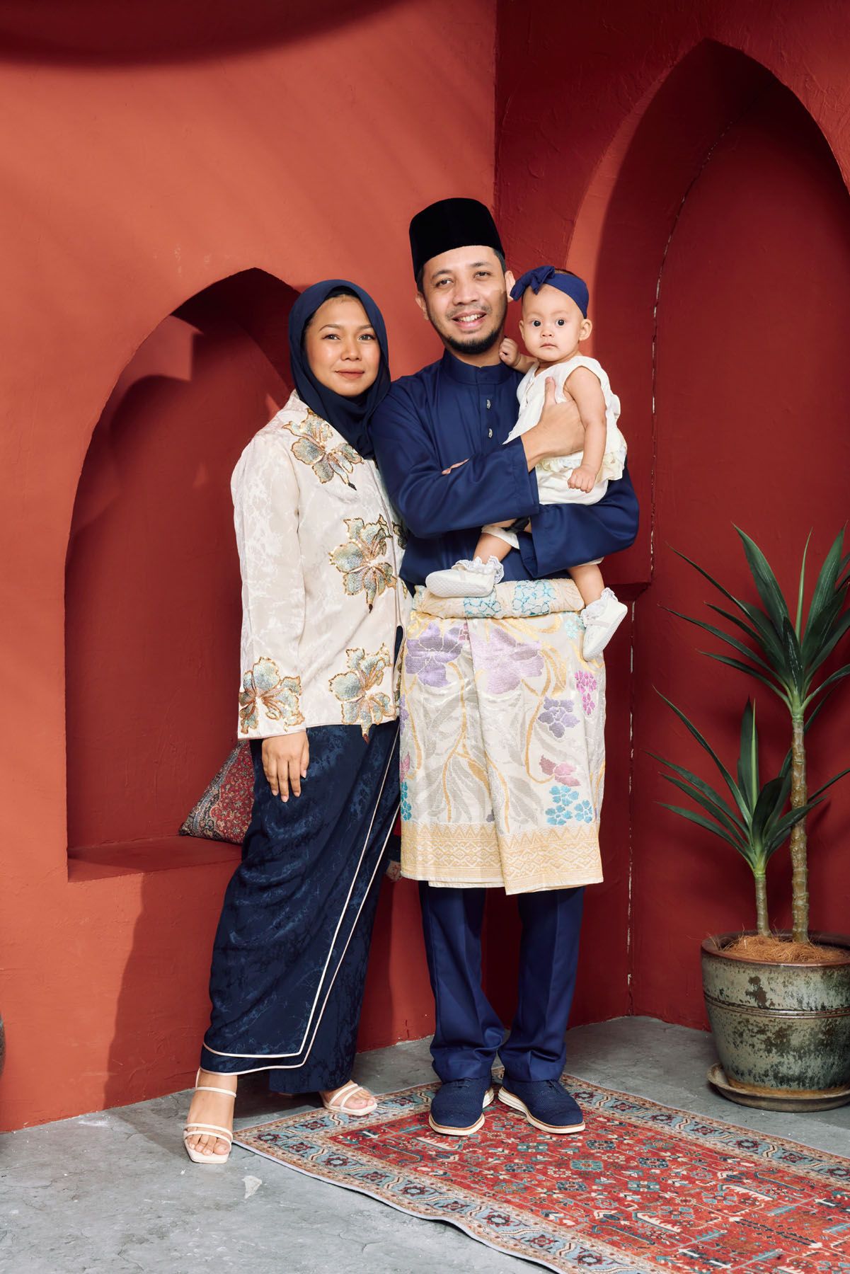 Young Malay family in navy blue traditional baju kurung and baju melayu posing against red arched wall.