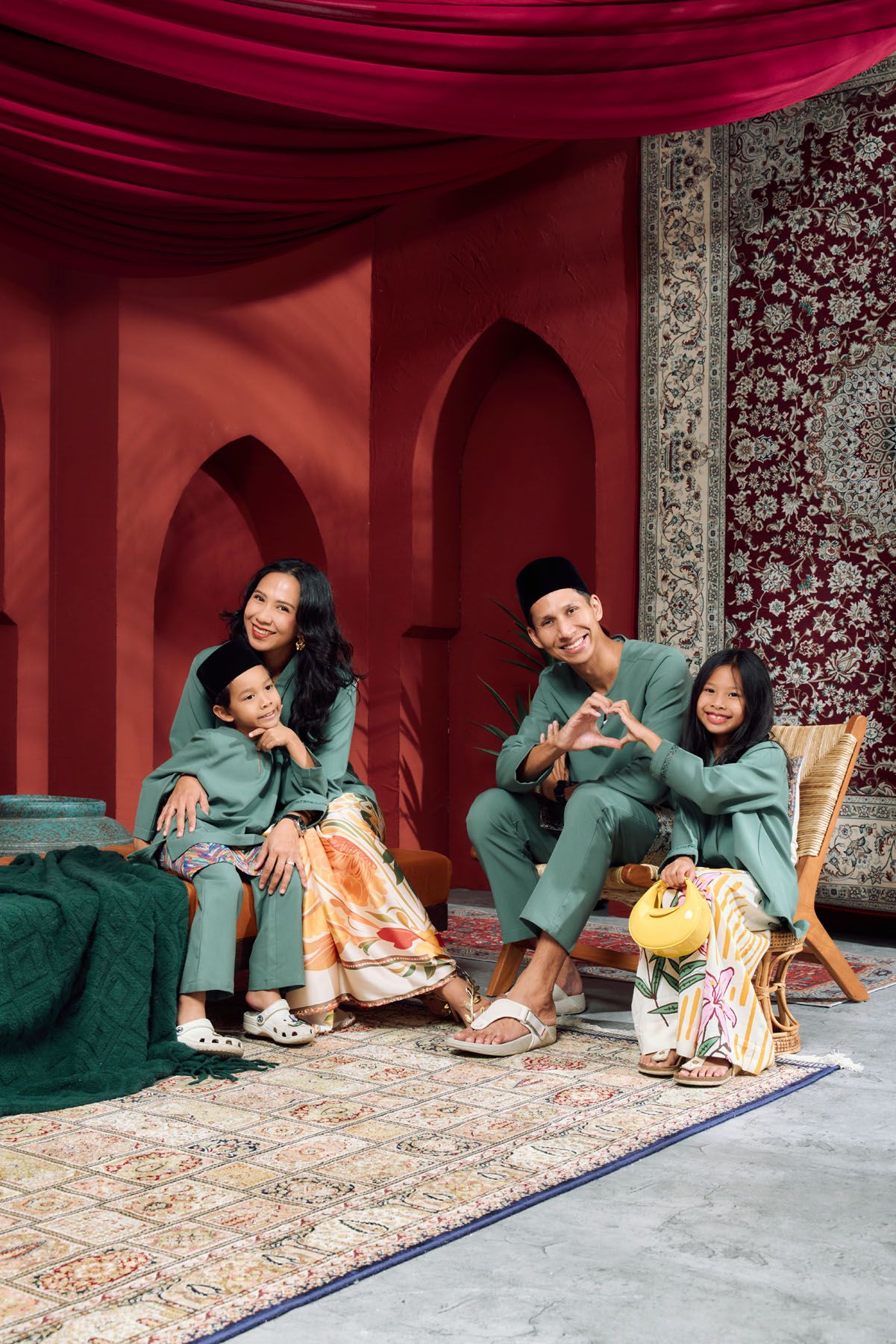 Family of four in green Baju Melayu and Baju Kurung posing playfully in a red Moroccan-inspired photo set.