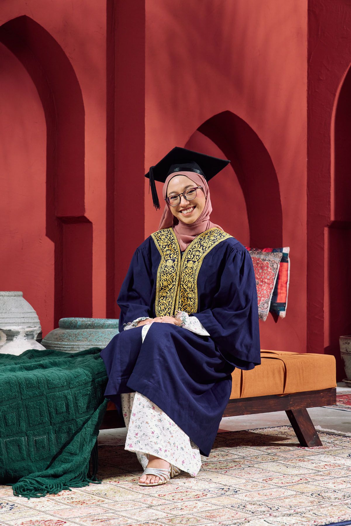 Smiling Muslim graduate in navy gown and hijab sits relaxed against a red Moroccan-style arched wall.