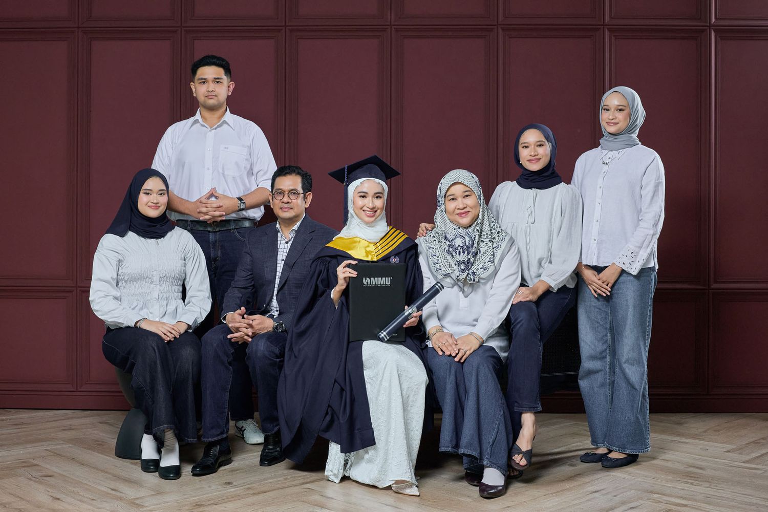 Female graduate in black cap and gown sits with family of seven against burgundy wall backdrop.