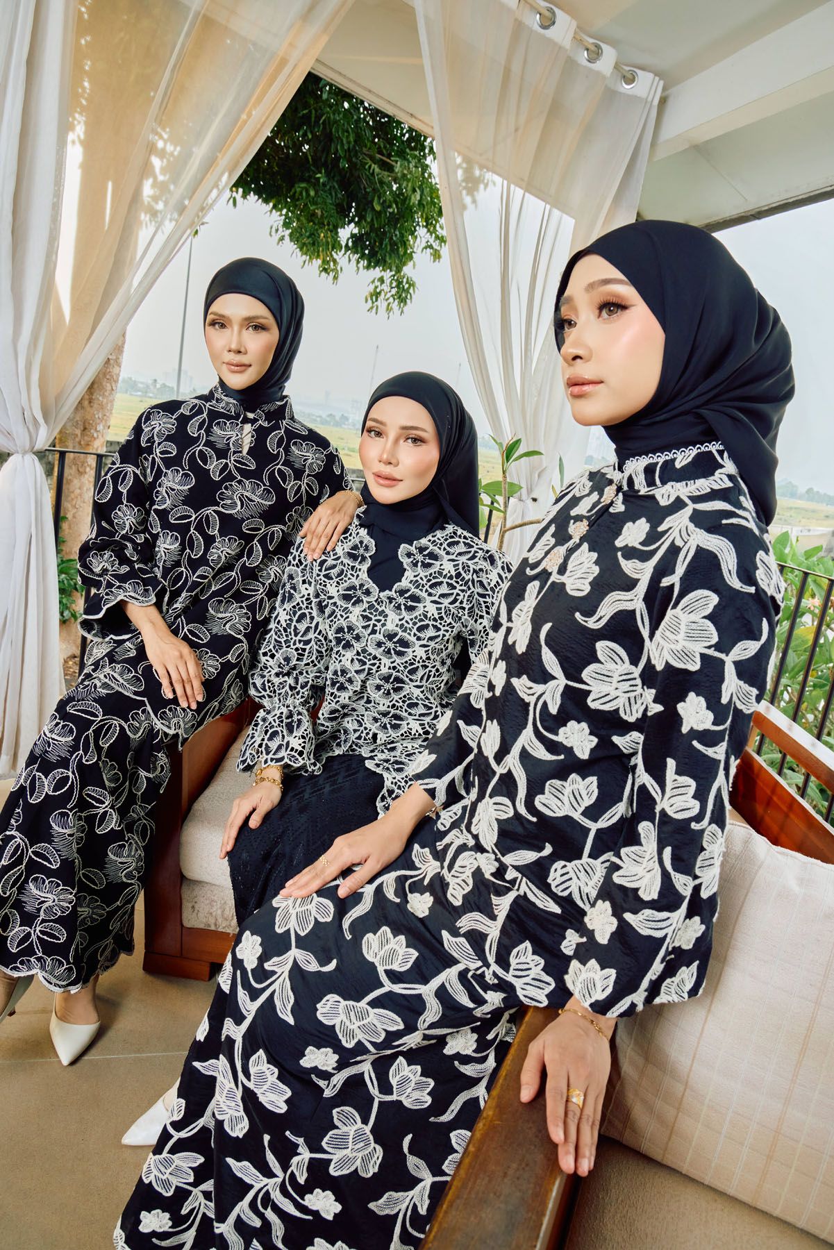 Three women in matching black floral baju kurung and hijabs pose elegantly indoors.