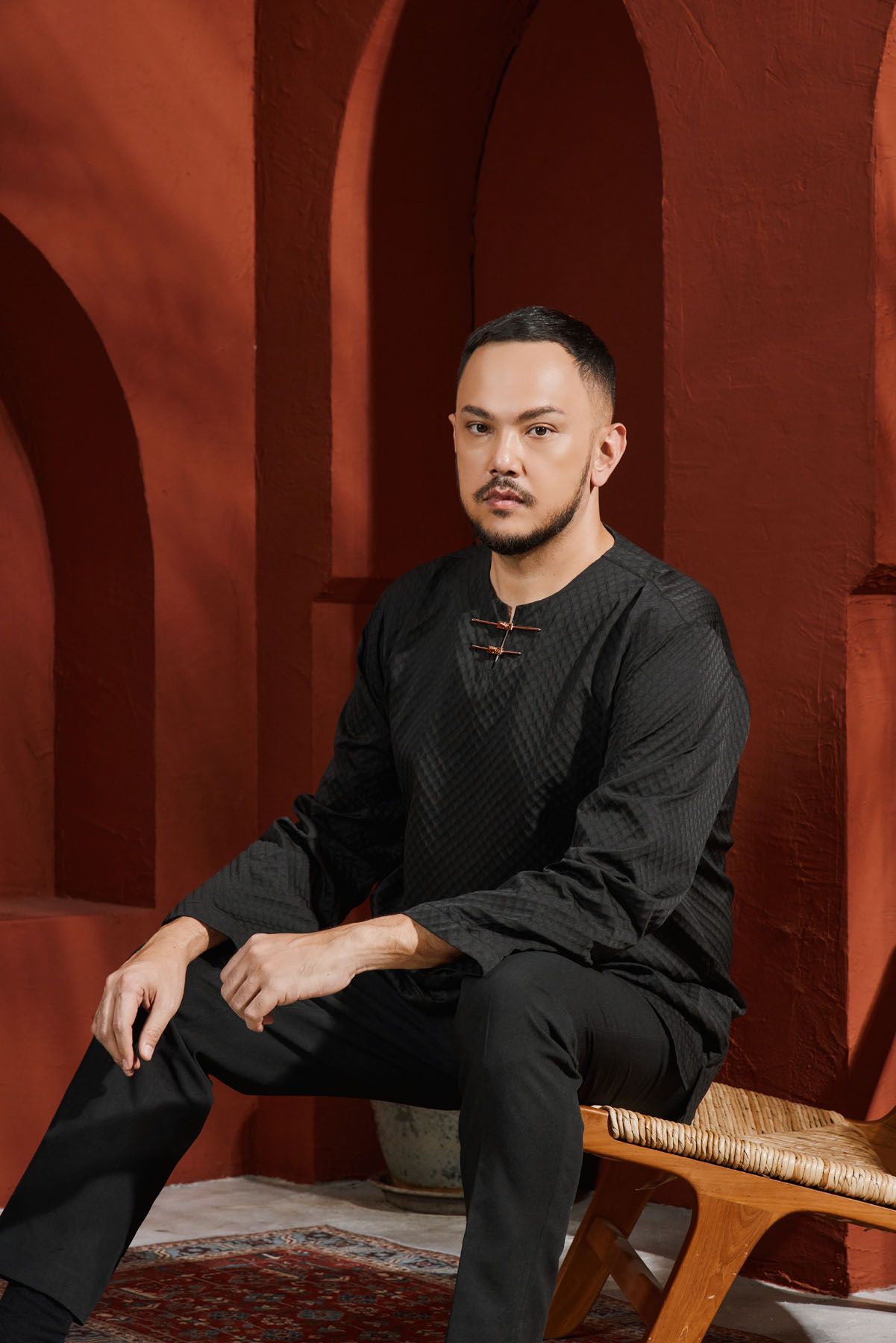 Man in black quilted baju melayu seated on a wooden bench against a red terracotta arched wall.