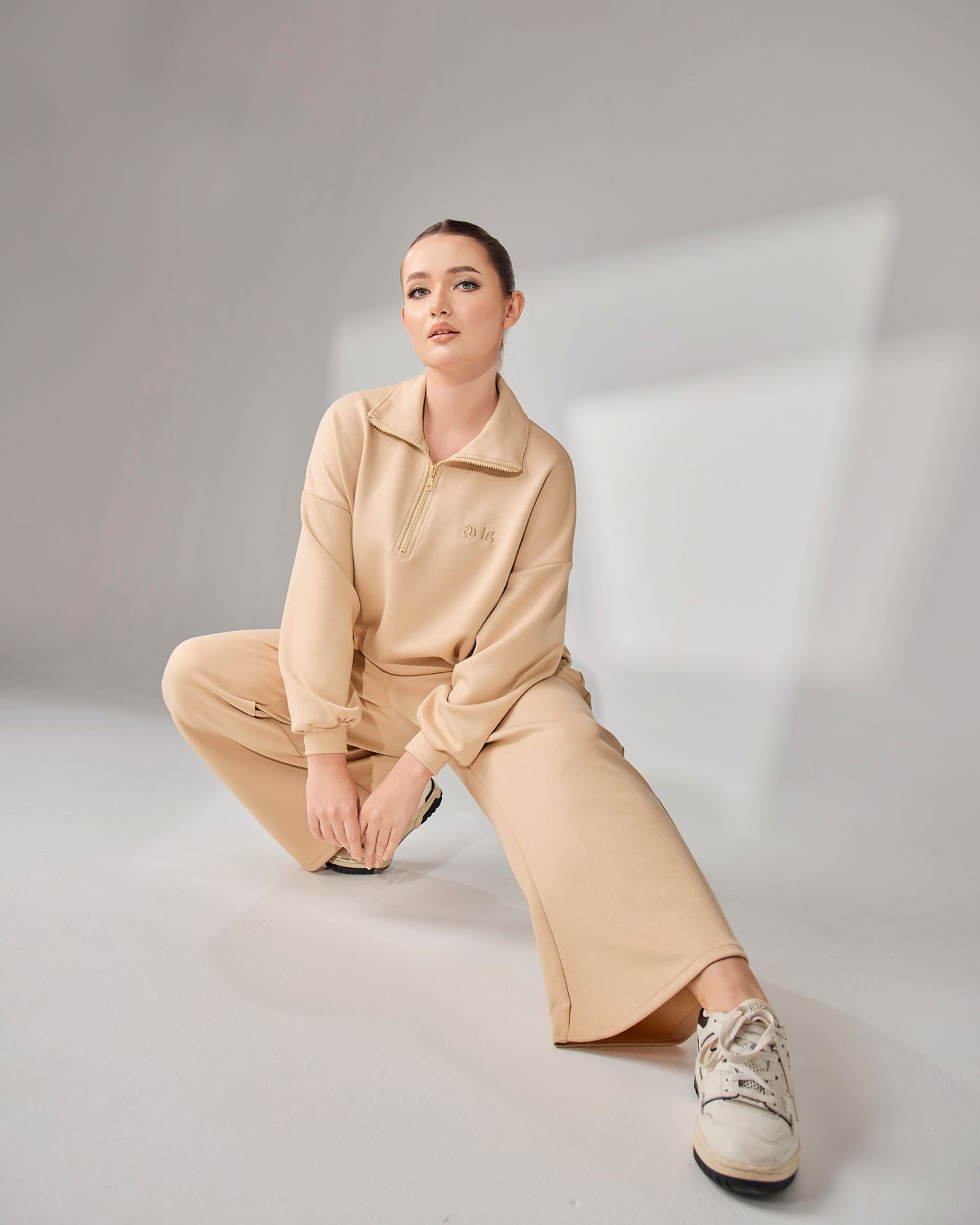 Woman in beige zip-up collared sweatsuit crouching gracefully in a bright minimalist studio setting.