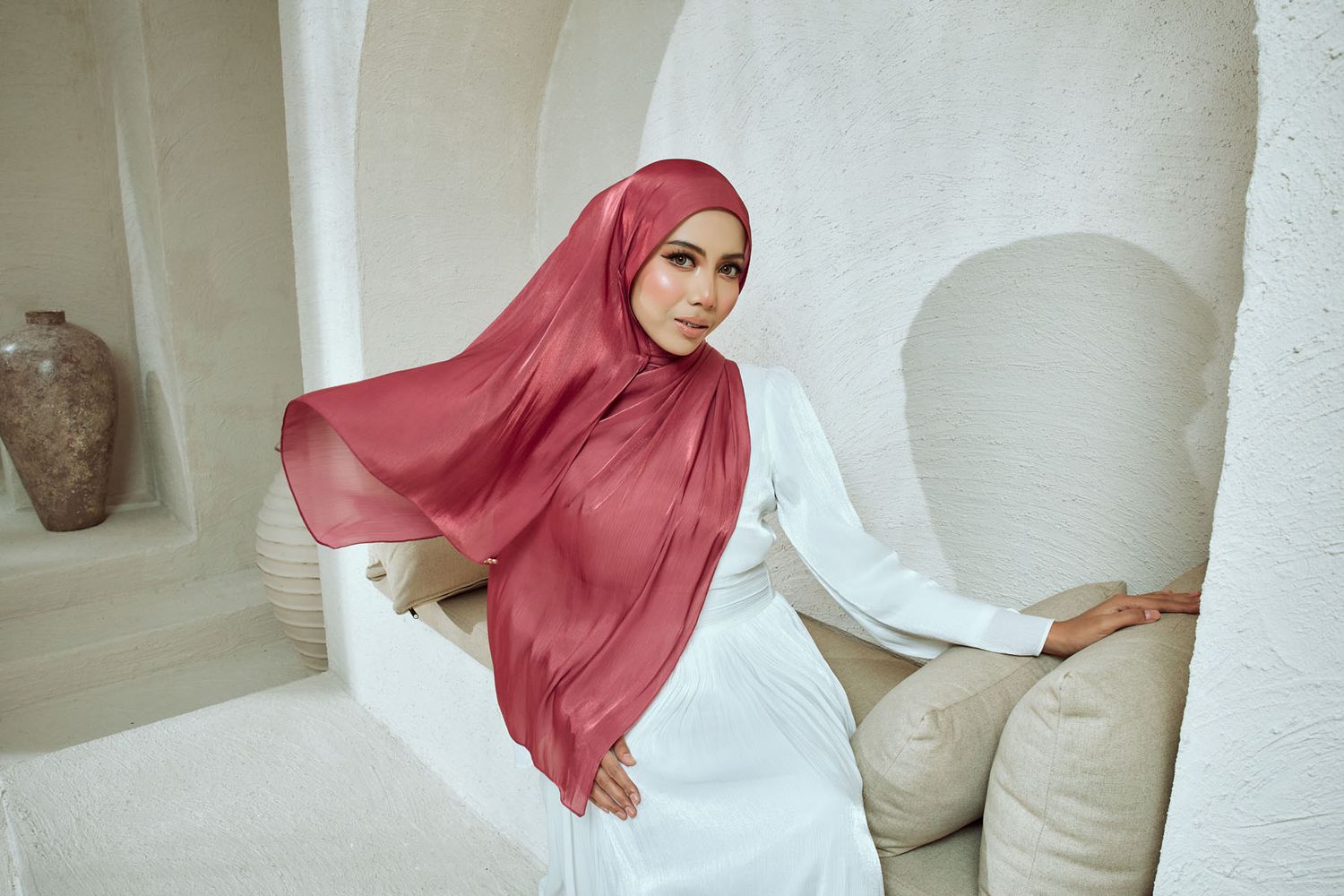Woman wearing a rose pink chiffon hijab with white dress seated in a whitewashed Mediterranean setting.