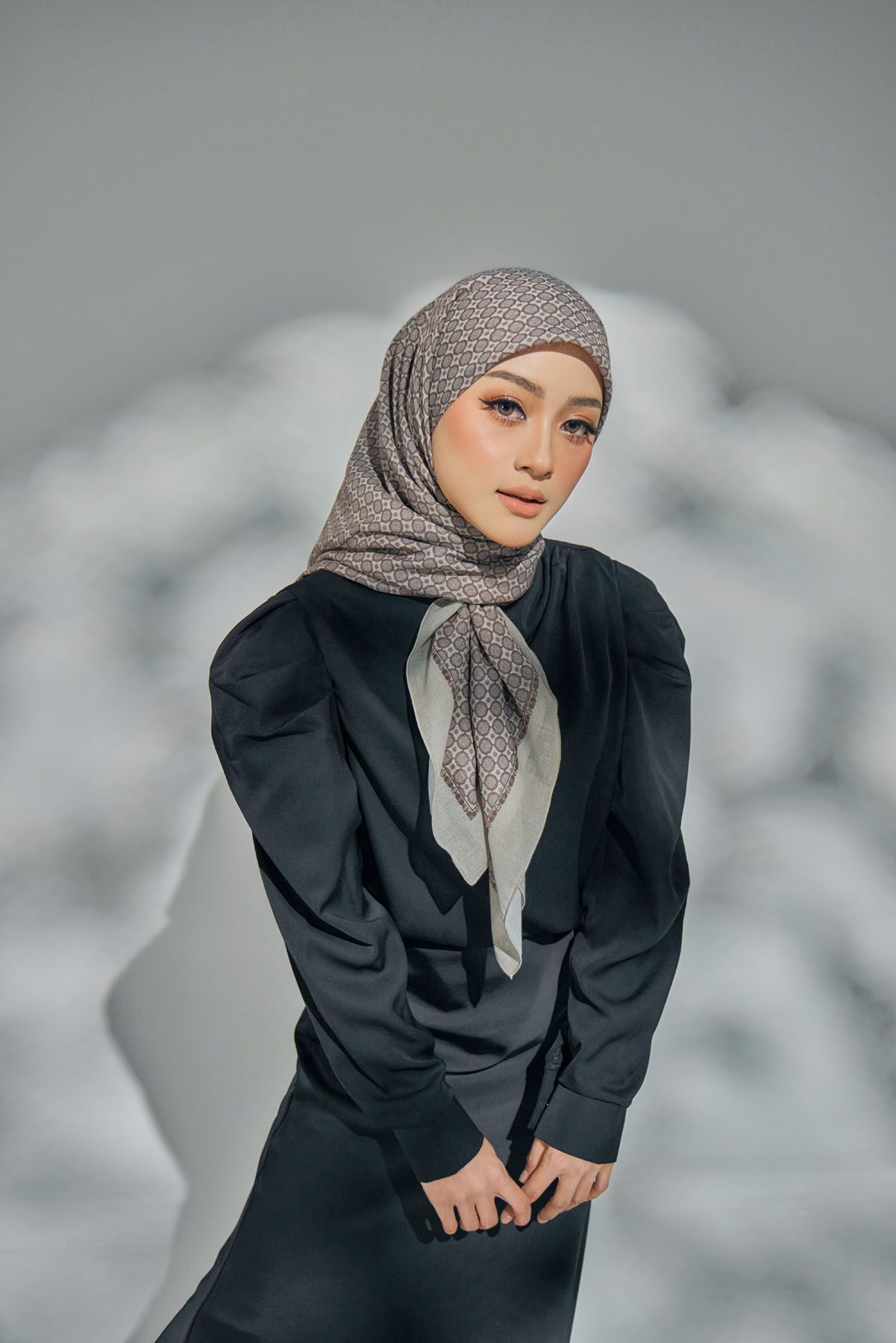 Woman in black blazer and grey patterned hijab posing confidently against soft grey background.