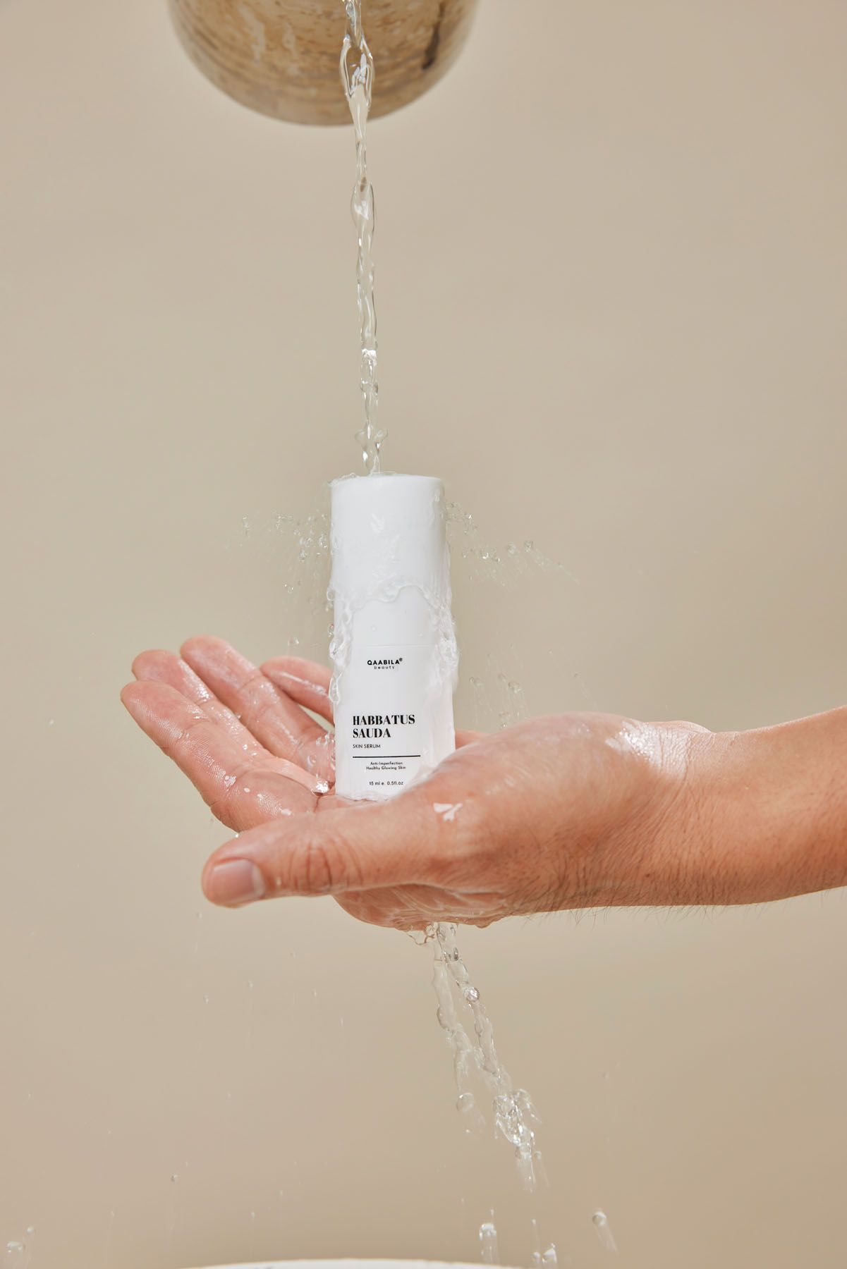 Water pouring over a hand holding a white skincare bottle labeled Barbatus Salda on beige background.