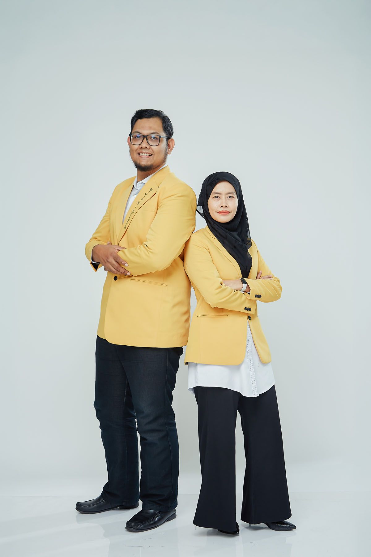 Asian man and Muslim woman in matching yellow blazers posing back-to-back with arms crossed.
