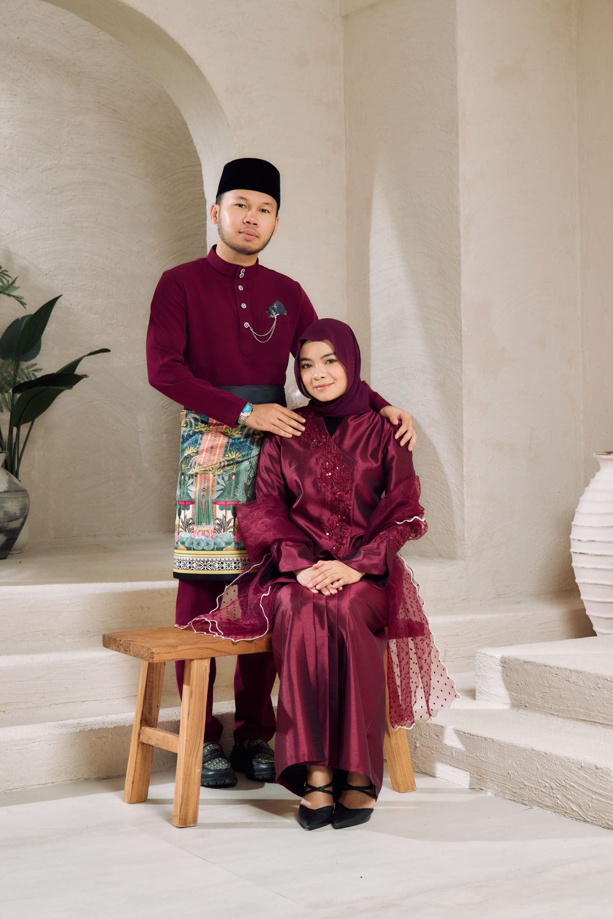 Malaysian couple in matching maroon Baju Melayu and Baju Kurung with batik accents in elegant interior setting.