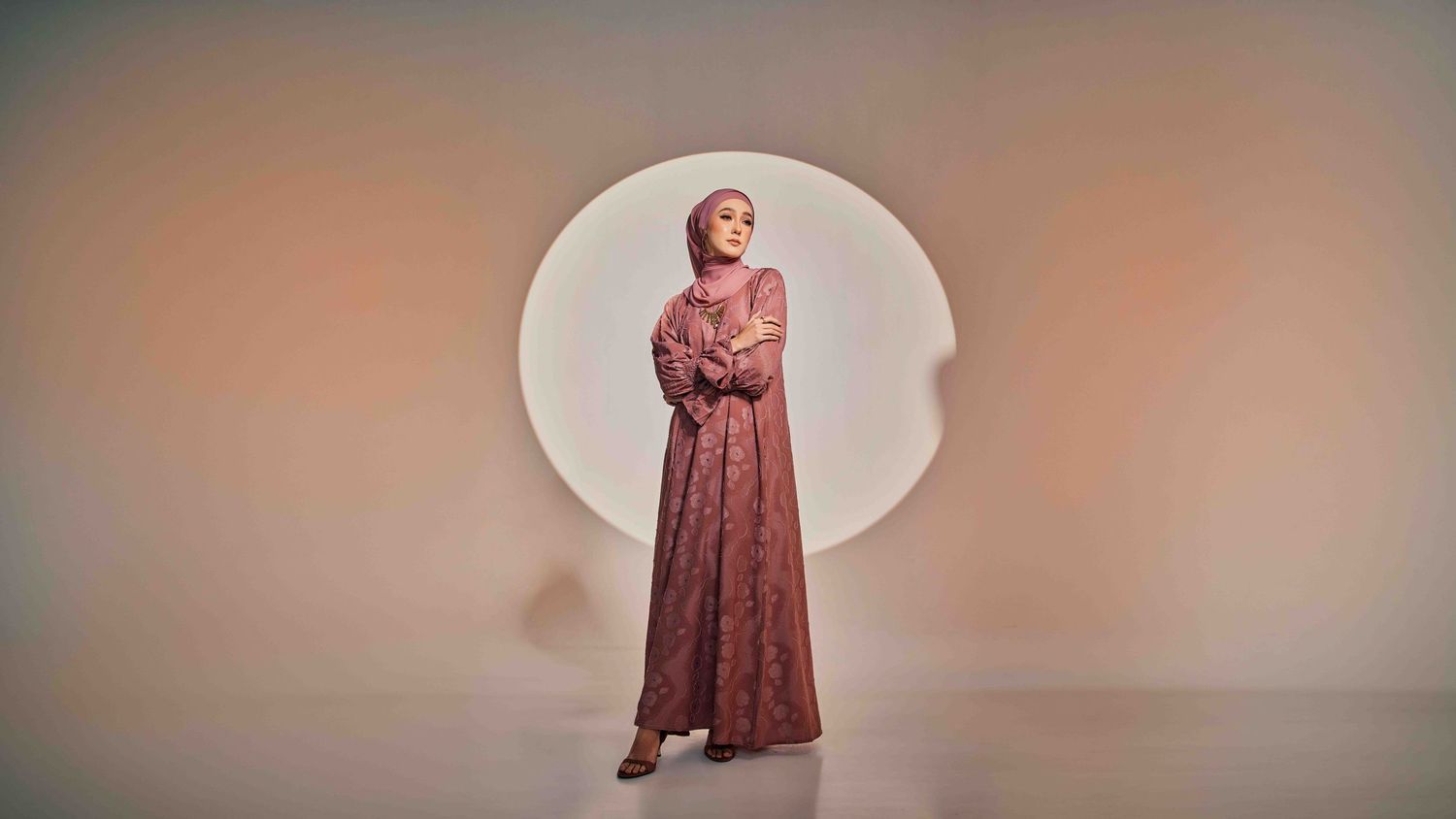 Muslim woman in dusty rose lace abaya and hijab standing before a glowing circular backdrop.