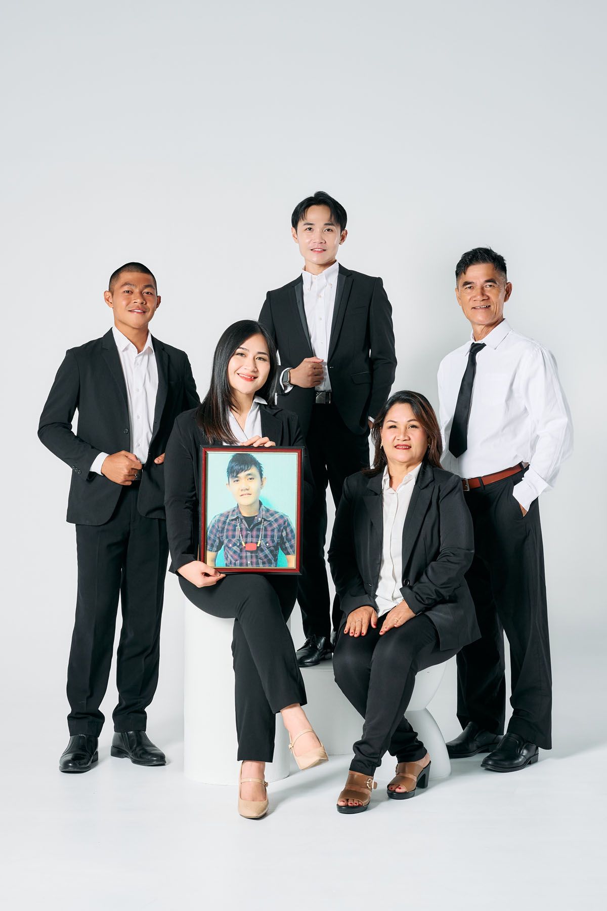 A family in black suits poses together, with a woman holding a framed portrait photo against a white background.