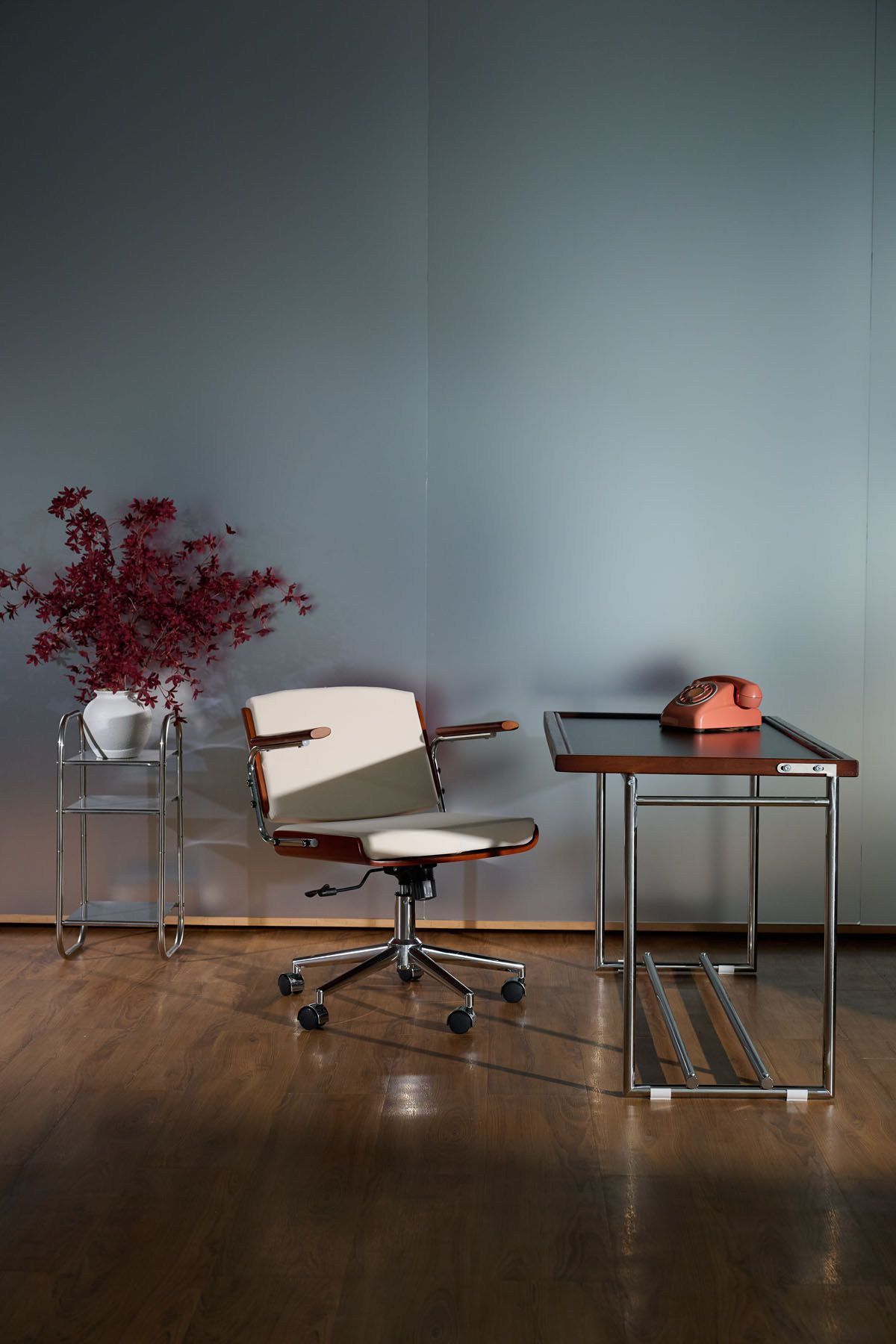Retro mid-century office setup with swivel chair, chrome side table, vintage orange rotary phone, and red floral arrangement on wood floor.