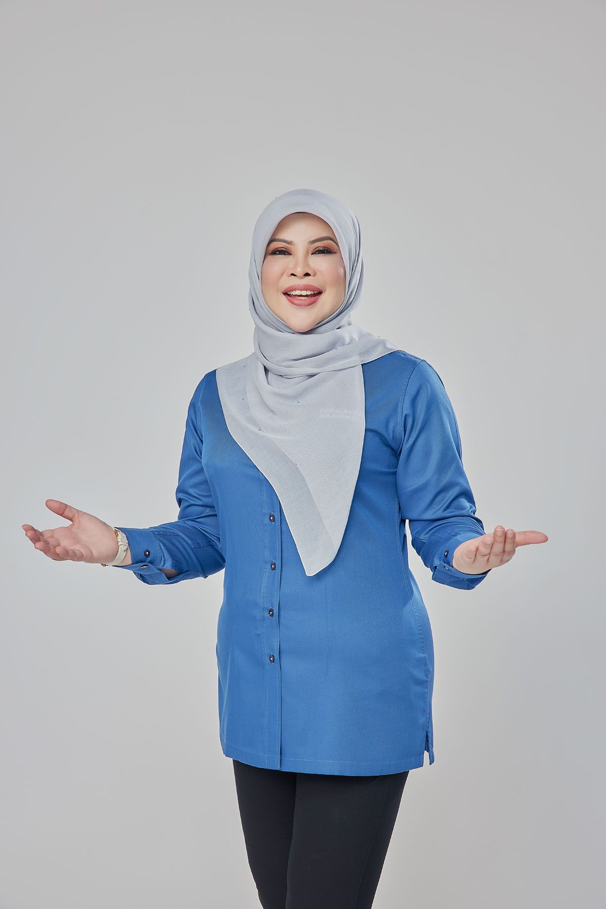 Smiling Muslim woman in blue tunic and grey hijab gesturing with open hands on grey background.