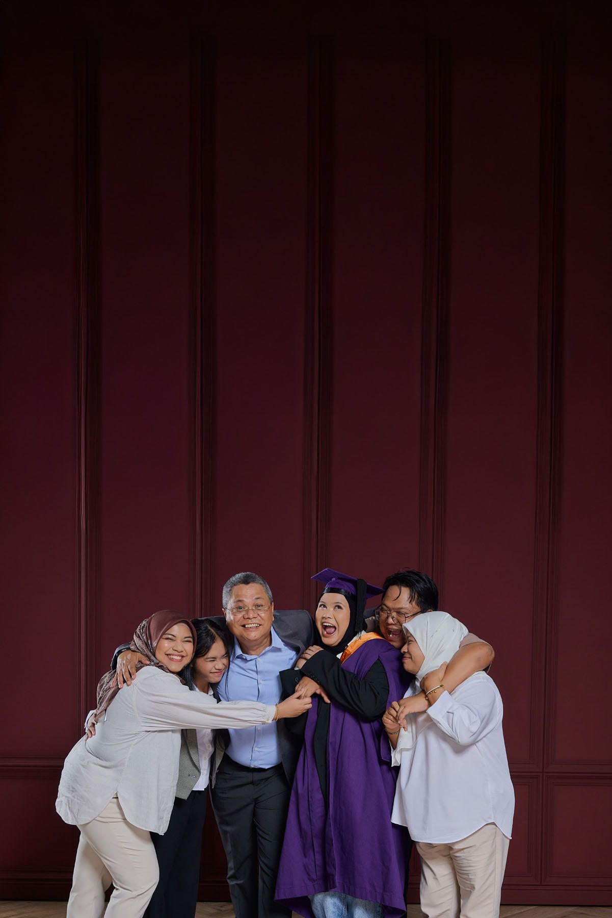 Joyful family hugging graduate in purple cap and gown against dark red paneled wall.