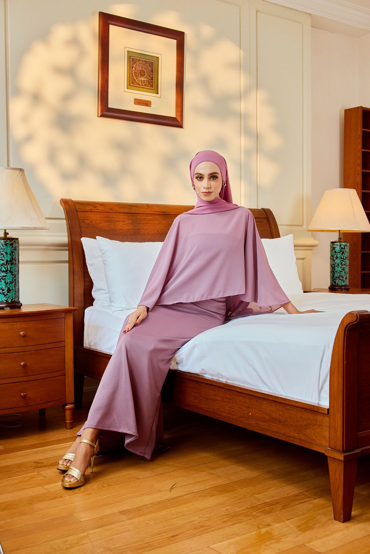 Woman in dusty pink batwing modest fashion top and wide-leg pants with matching hijab on wooden bed.
