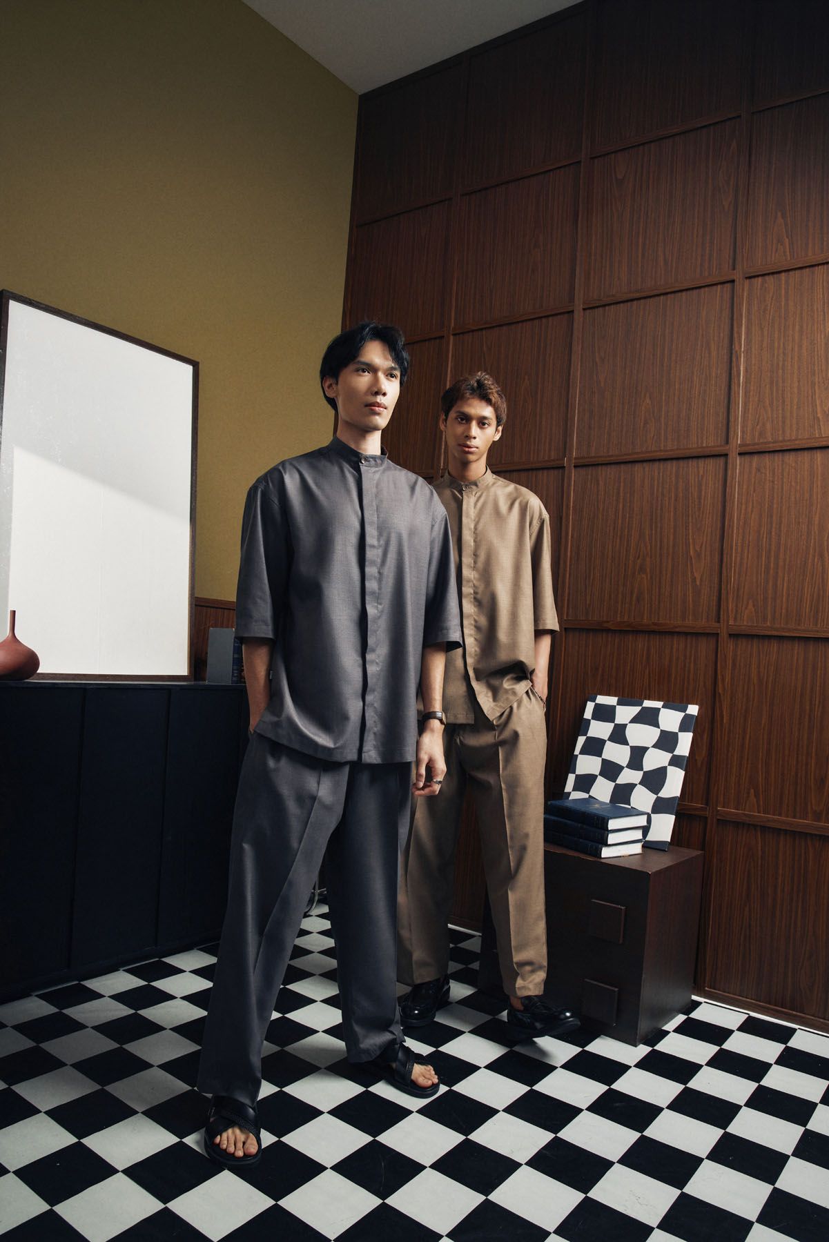 Two men in gray and tan traditional matching sets stand on checkered floor in modern interior.