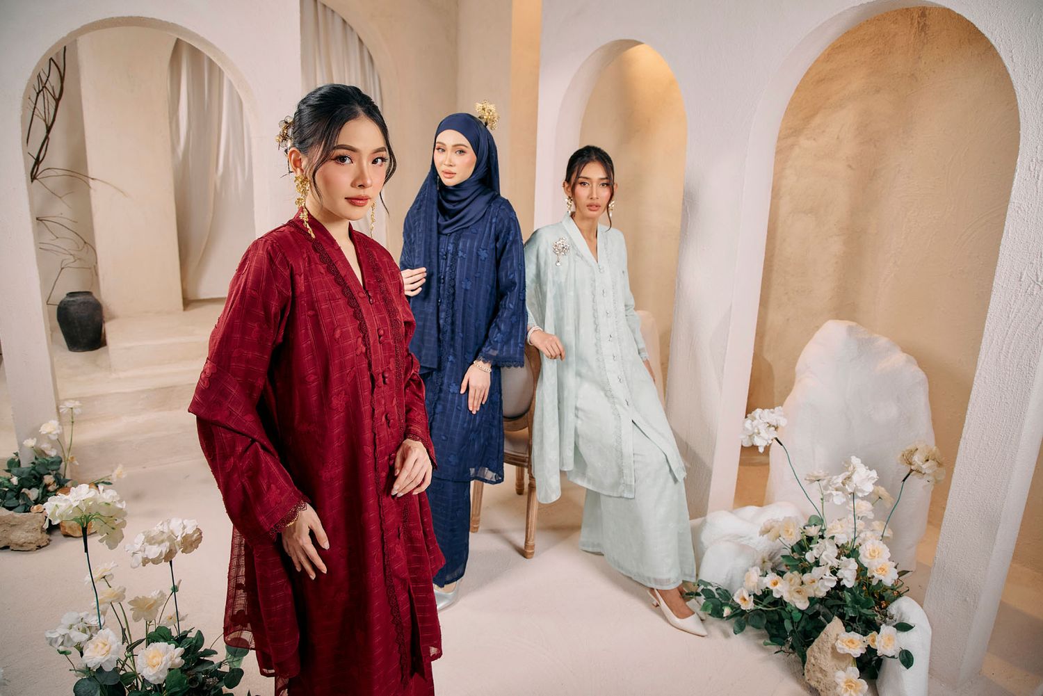 Three models wear elegant long tunics in burgundy, navy, and mint green hijab styles in a floral decorated studio.