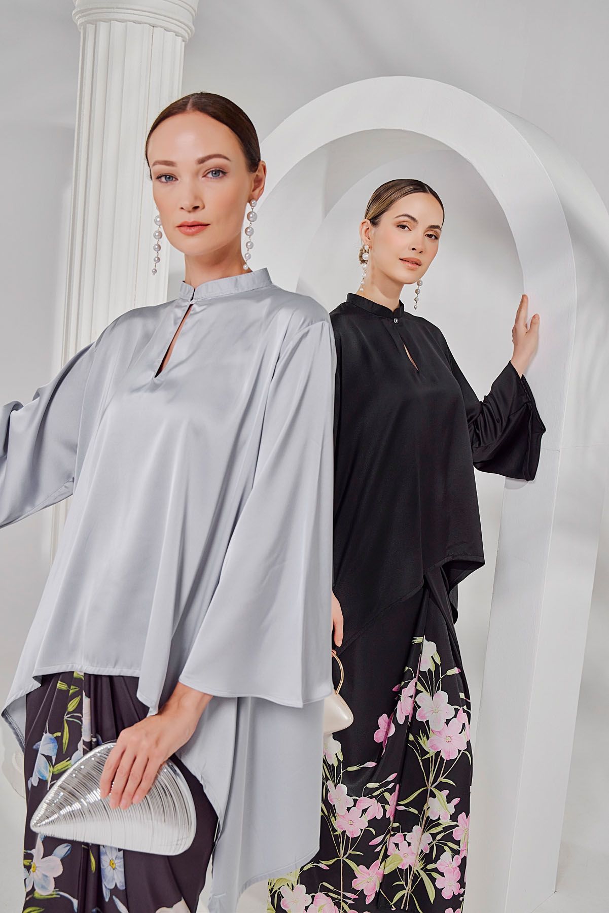 Two women model elegant satin kaftan tops in silver and black with floral skirts near white arches.