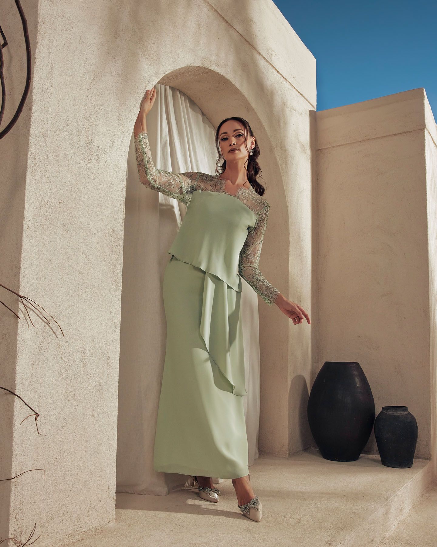 Woman in sage green lace peplum top and maxi skirt stands in circular stone archway under blue sky.