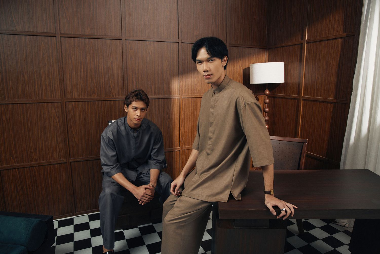 Two young men in modern baju melayu outfits pose in a sophisticated wood-paneled room.