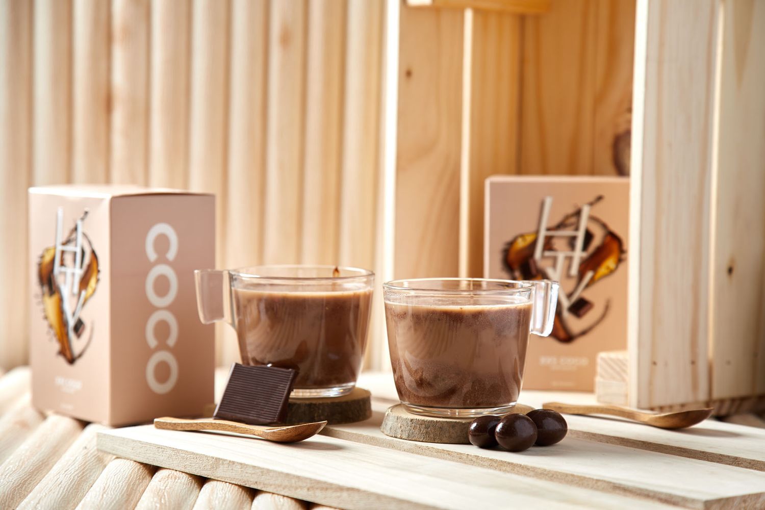 Two glass cups of Coco hot chocolate with chocolate pieces on wooden coasters beside product boxes.