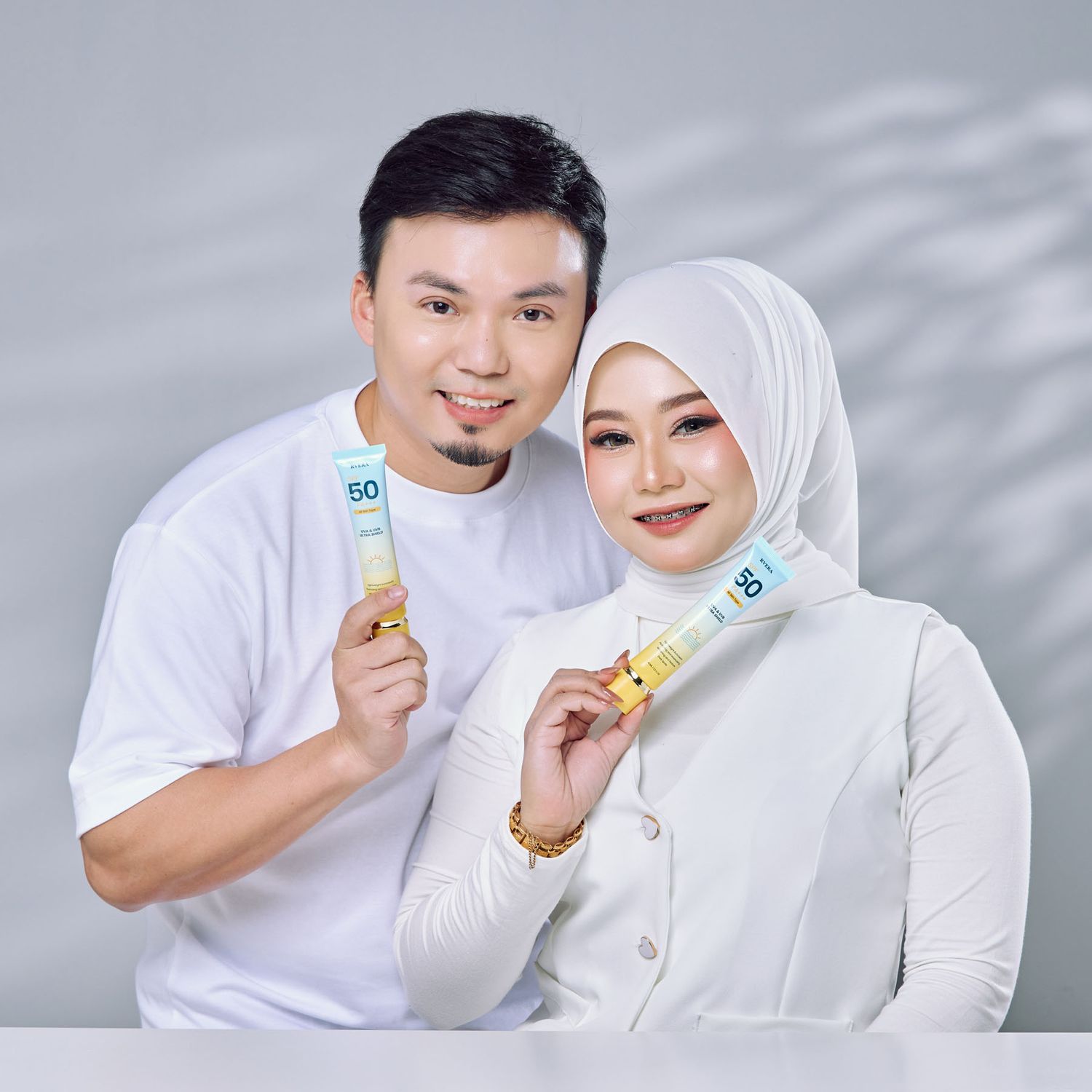 Couple in white outfits holding SPF50 sunscreen tubes together in a bright studio photo setting.