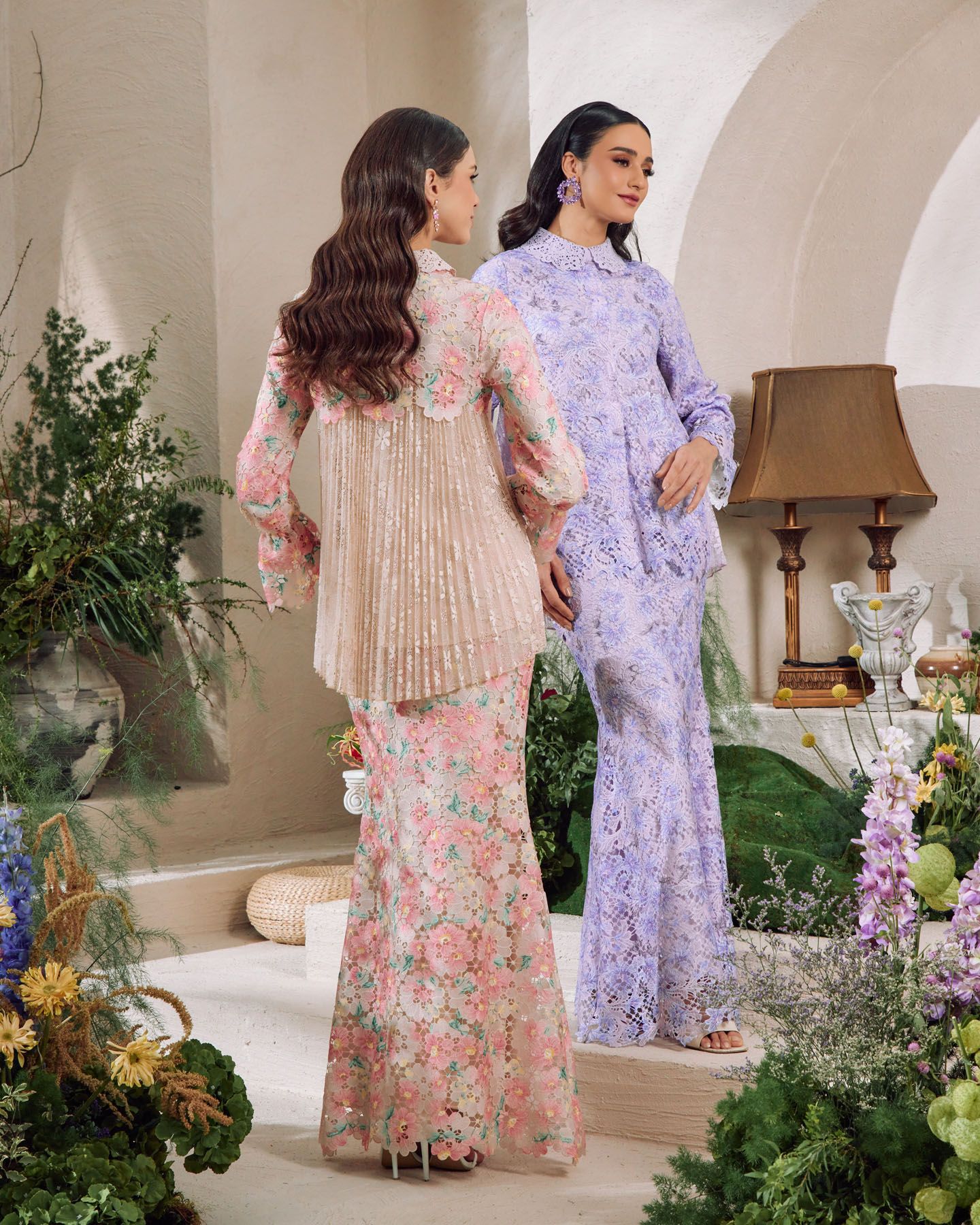 Two models showcase ornate Malaysian baju kurung, one in pink floral lace and one in lavender lace, among garden blooms.