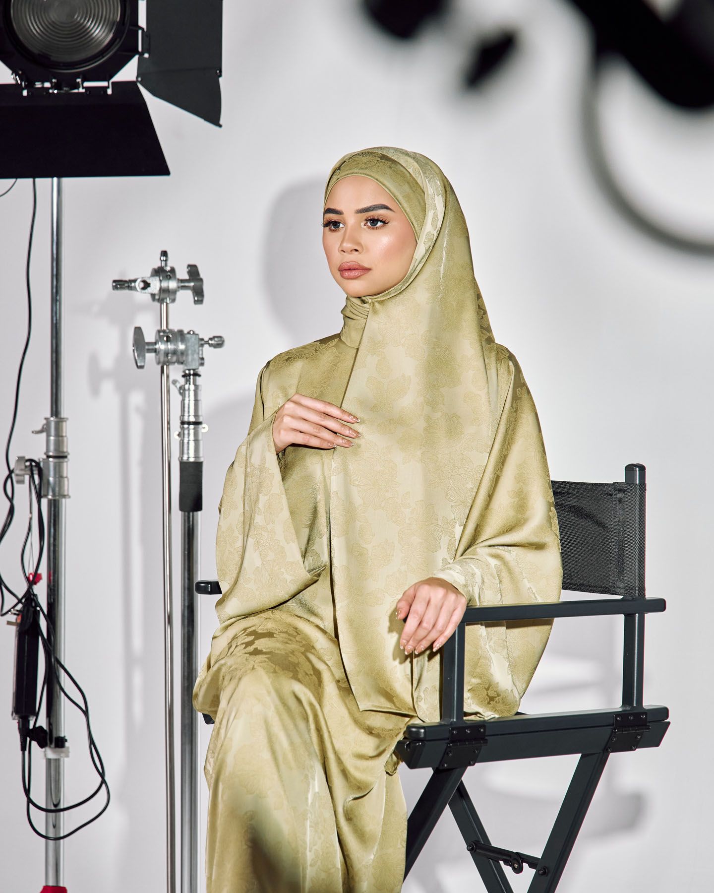 Woman in olive green satin hijab and matching outfit sits in director chair.