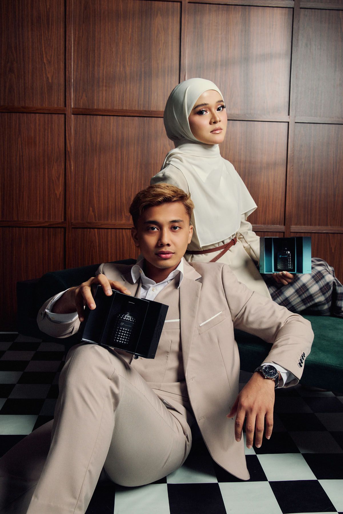 Elegant couple in matching beige suits posing with black cosmetic compacts in a sophisticated interior.