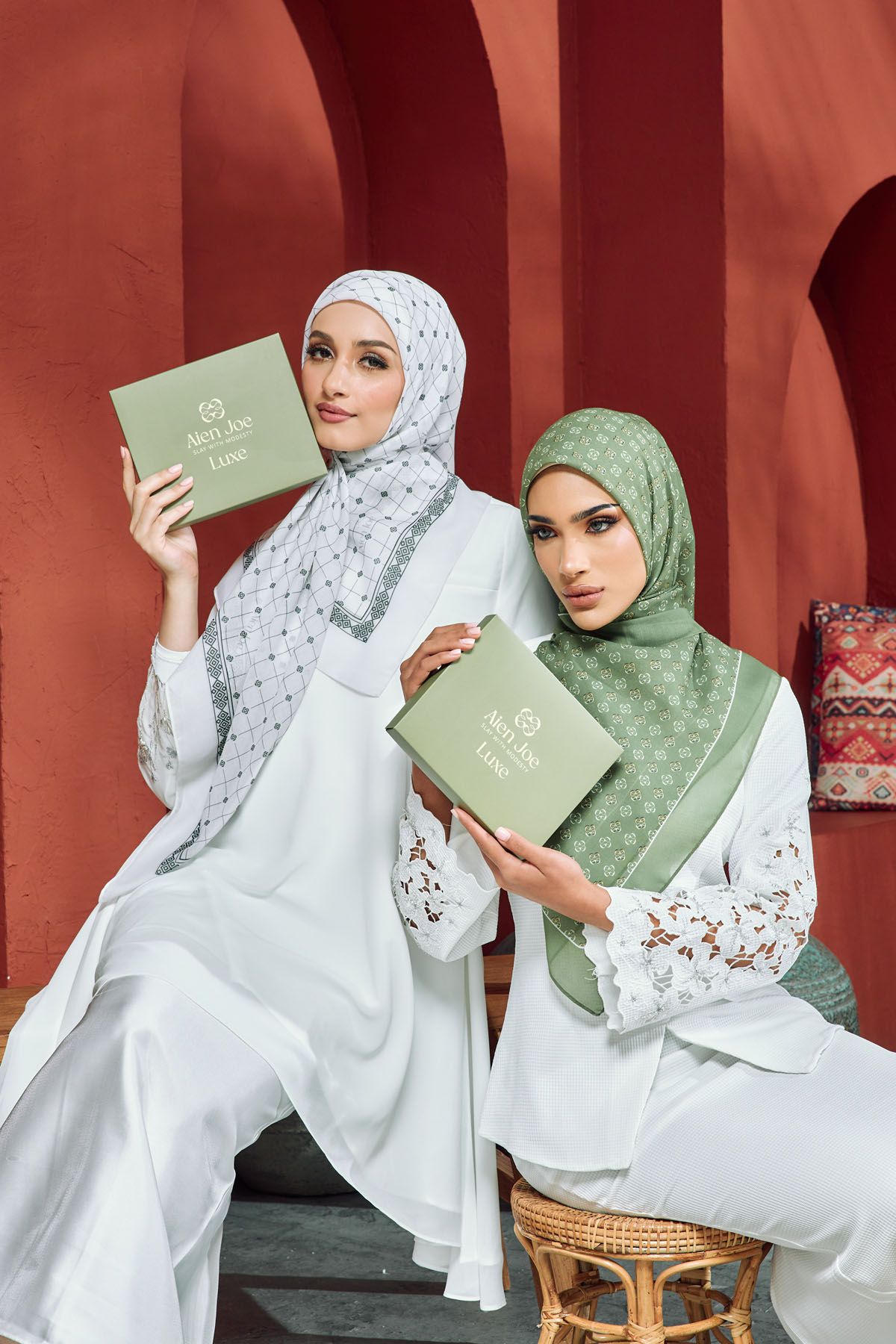 Two women in white modest outfits holding green gift boxes while wearing hijabs.