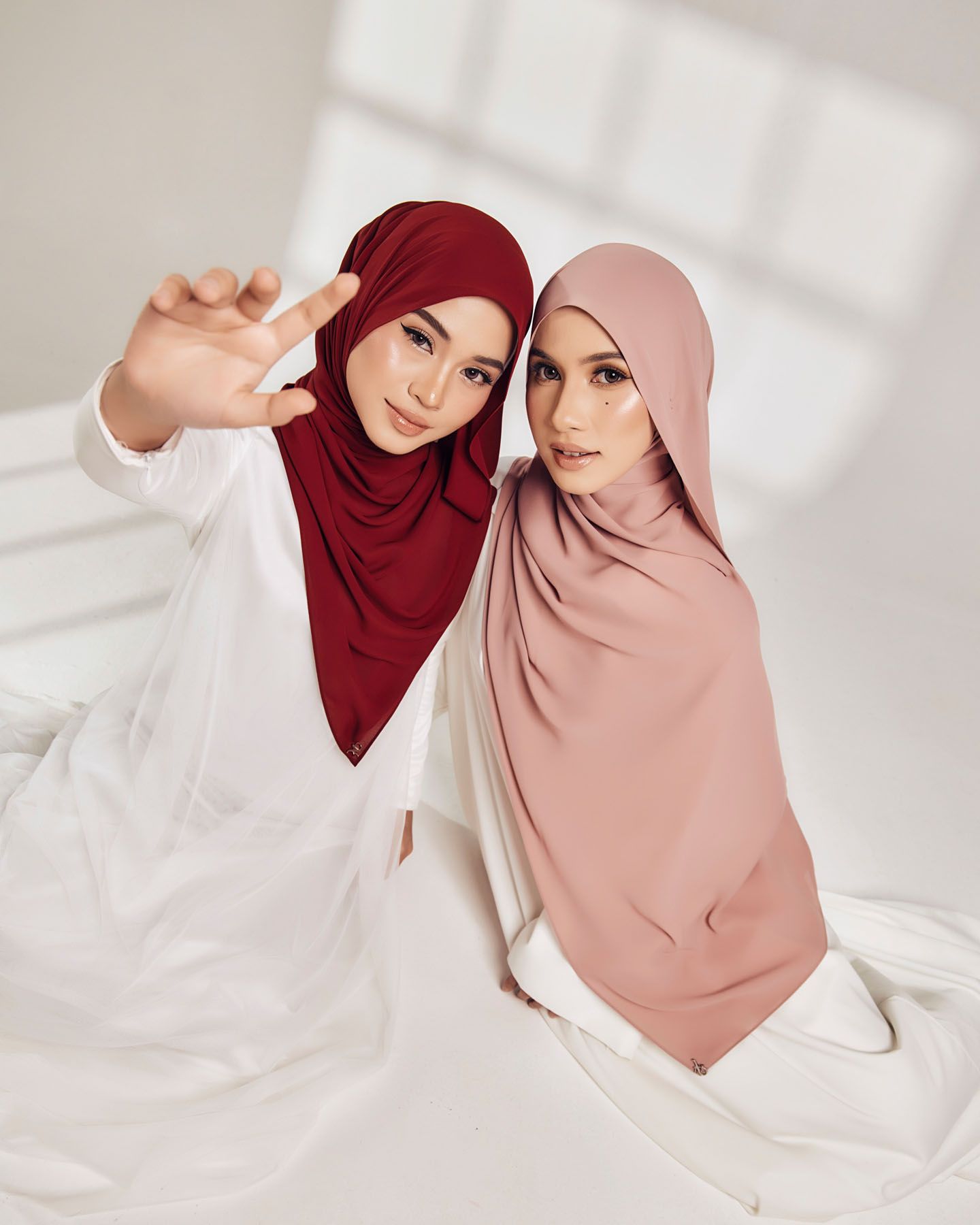 Two women wearing colorful hijabs in red and pink pose together in a bright white studio setting.