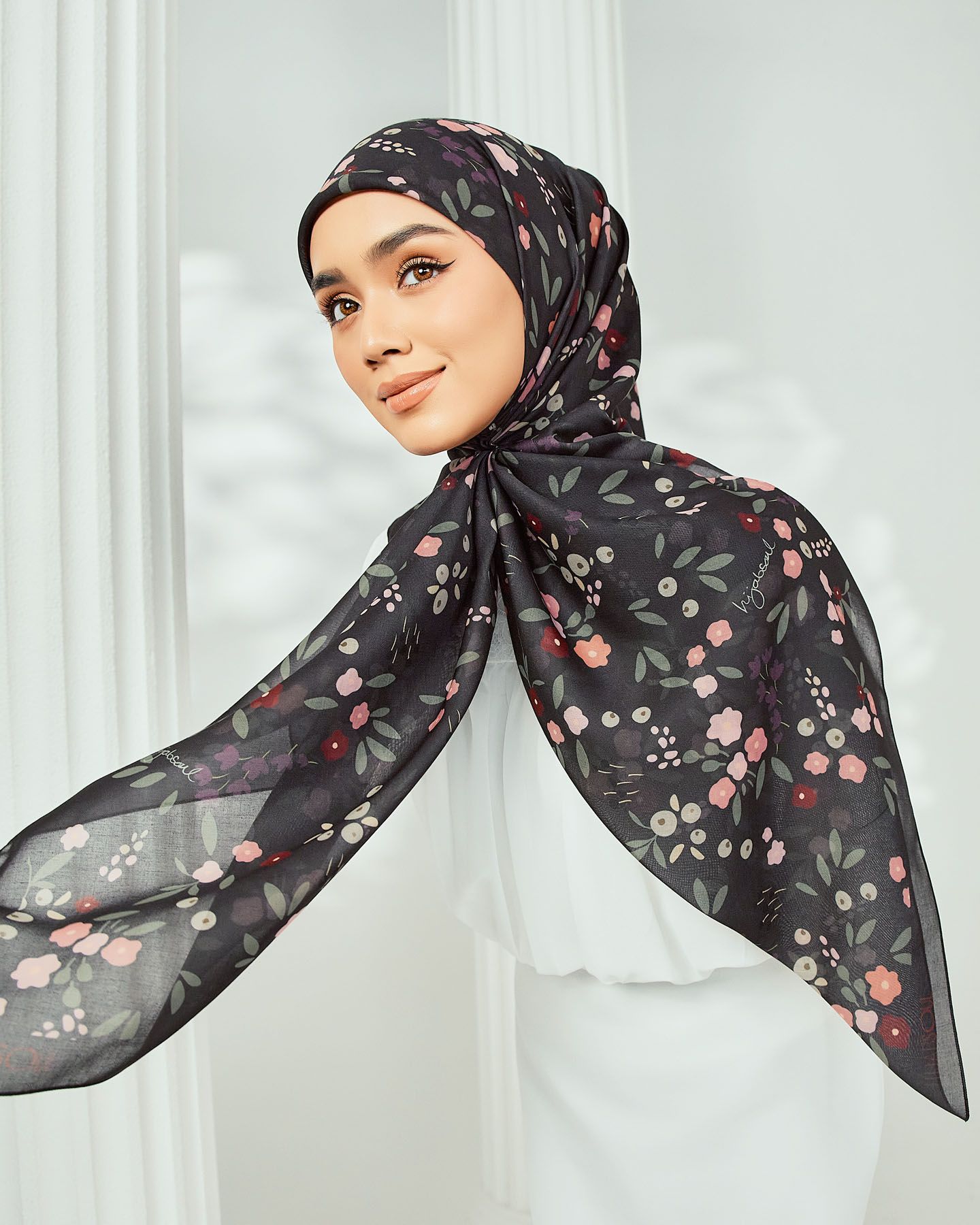 Woman modeling a black chiffon hijab with delicate pink floral print in an elegant pose.