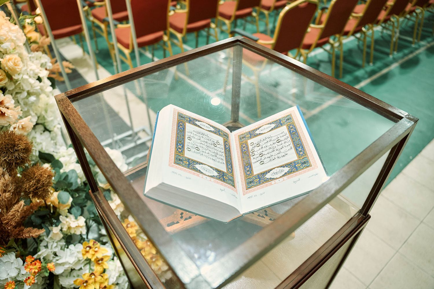 Open Quran displayed on glass stand surrounded by white floral arrangements at Islamic ceremony.