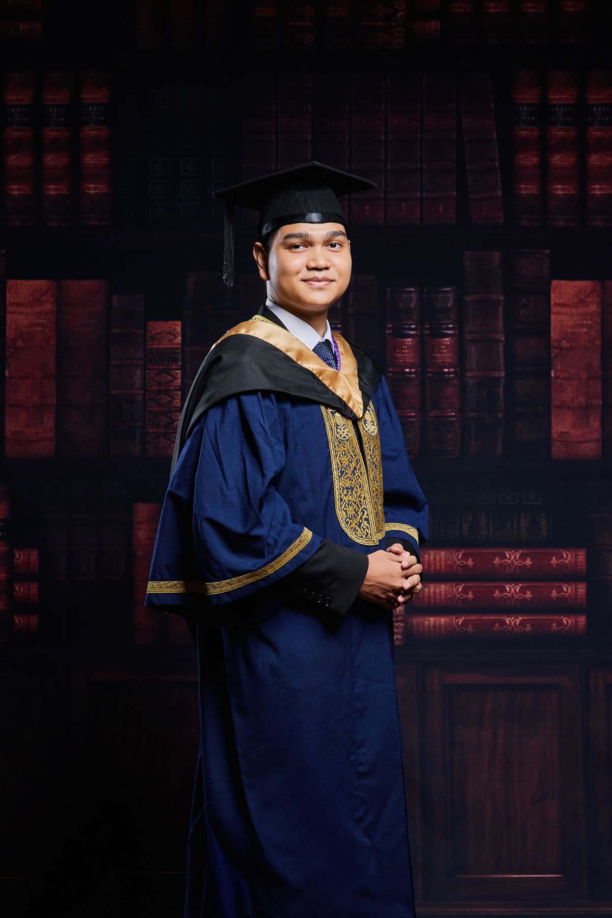 Young man in blue and gold graduation gown and mortarboard cap posing formally against dark bookshelf background.
